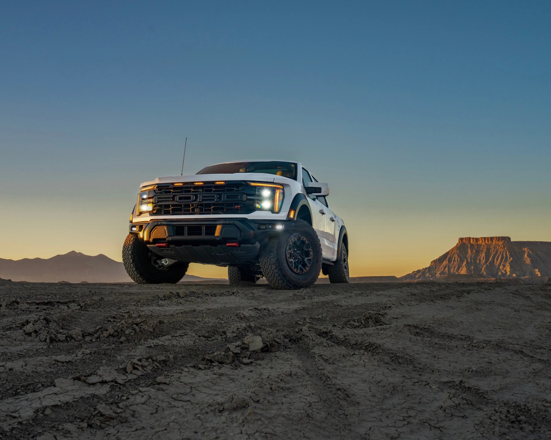 White Ford off-road truck with headlights on parked on dirt with mountains and sunset sky in background.