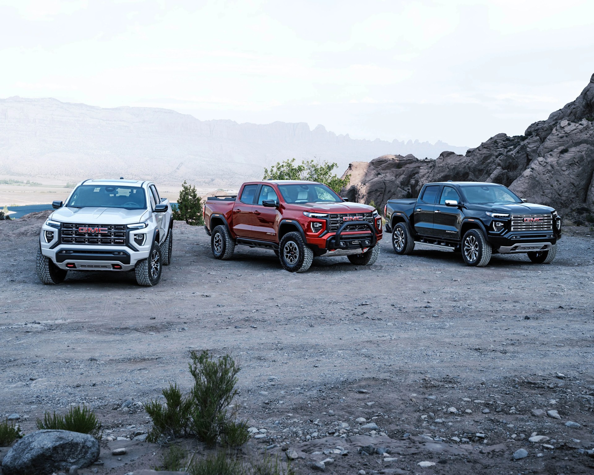 Three GMC pickup trucks in white, red, and black parked on rocky terrain with mountains in the background.