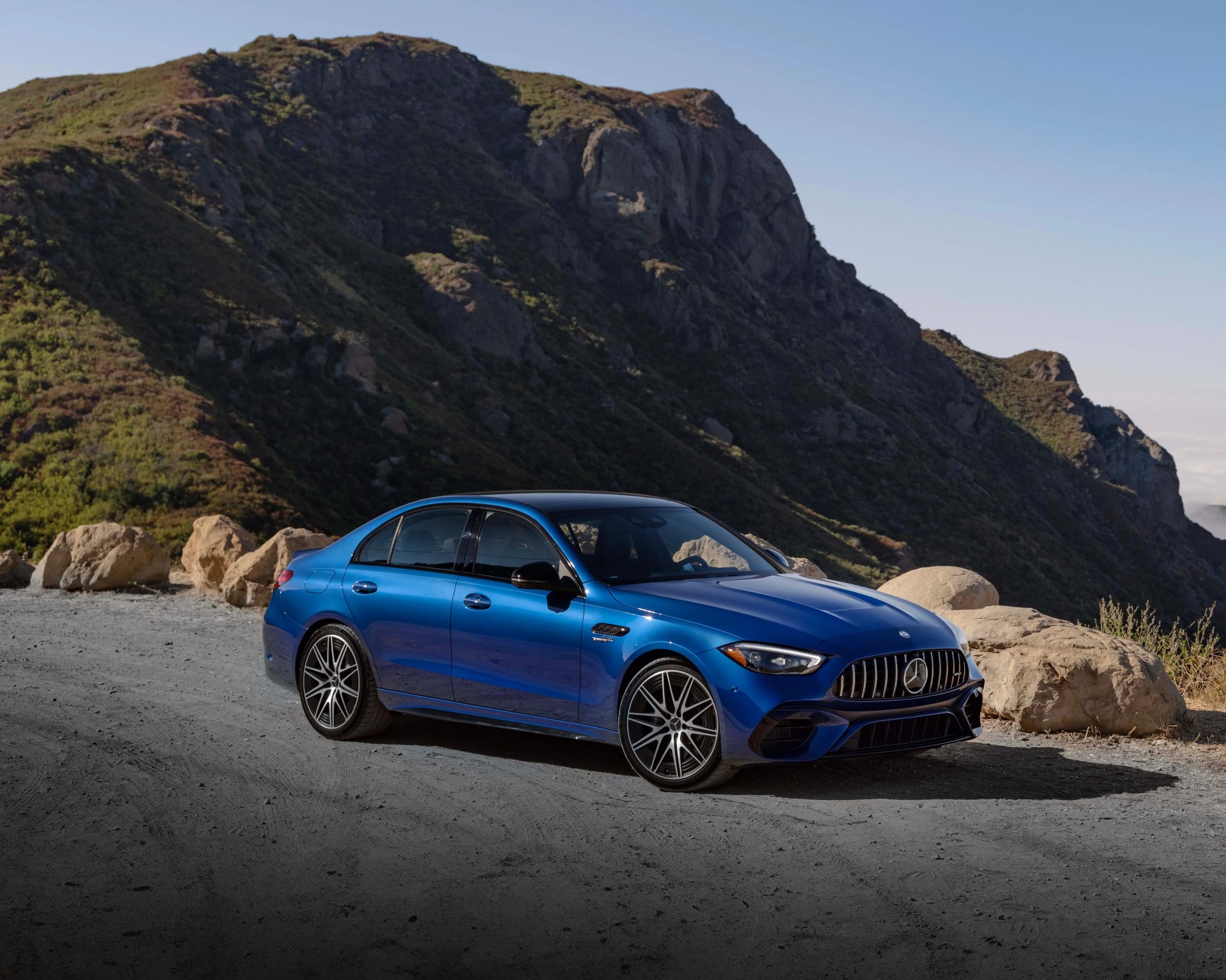 Blue Mercedes-Benz AMG sedan parked on a dirt road with rocky hills in the background.