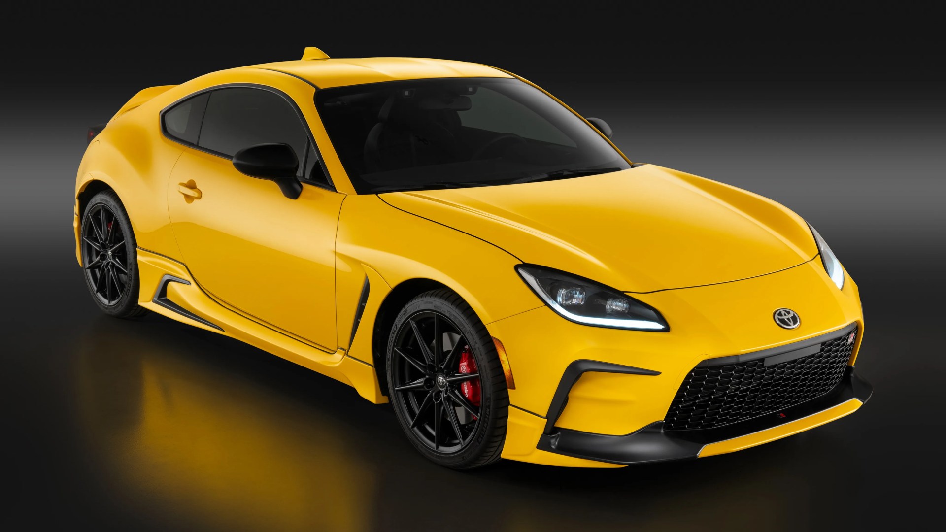 Yellow Toyota sports coupe with black wheels and red brake calipers on a dark reflective surface.