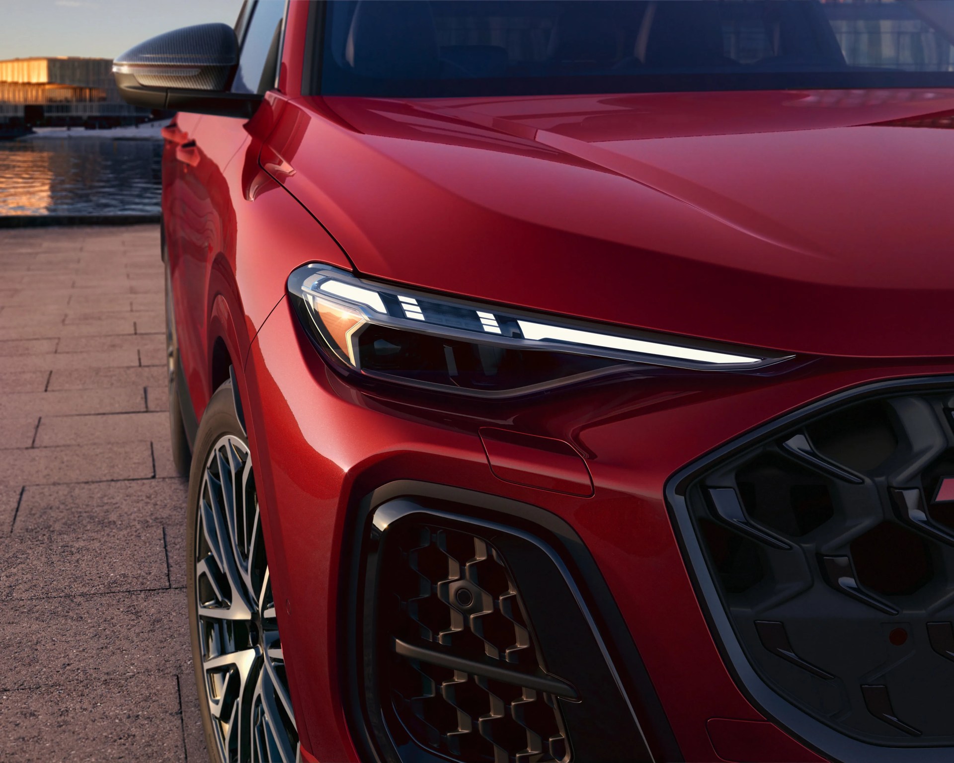 Front left side of a red car showing a sleek LED headlight, black honeycomb grille, and alloy wheel on a paved surface near water.