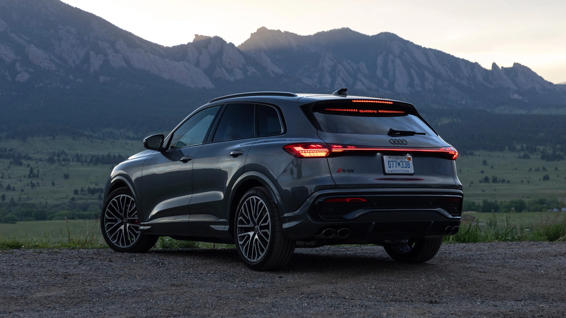 Dark gray Audi SQ5 SUV with illuminated rear lights parked on gravel with mountains in the background at dusk.