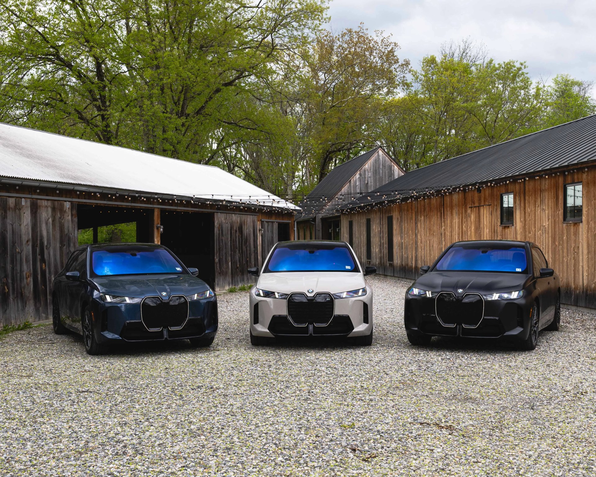 Three BMW iX electric SUVs in black, beige, and dark blue parked on gravel between rustic wooden buildings with string lights overhead.
