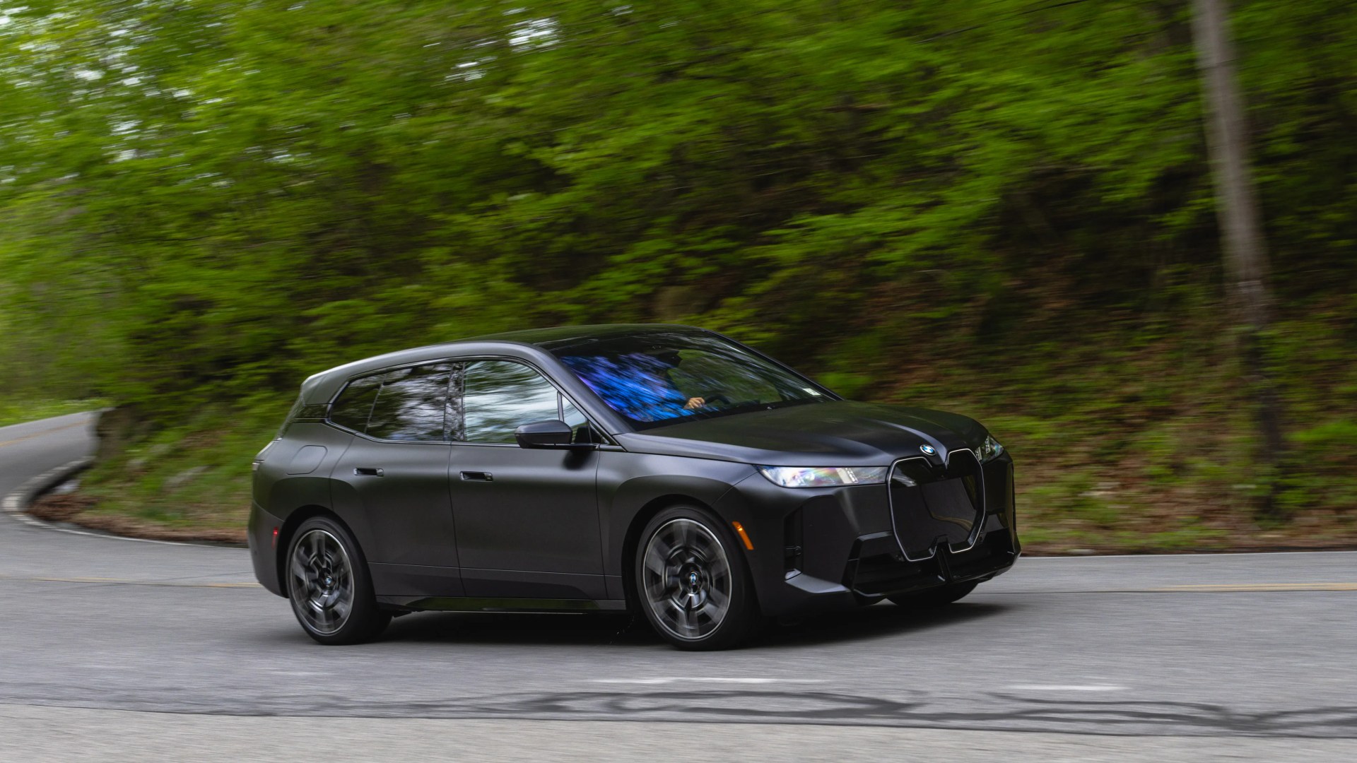 Matte black BMW SUV driving on a winding road with green trees in the background.