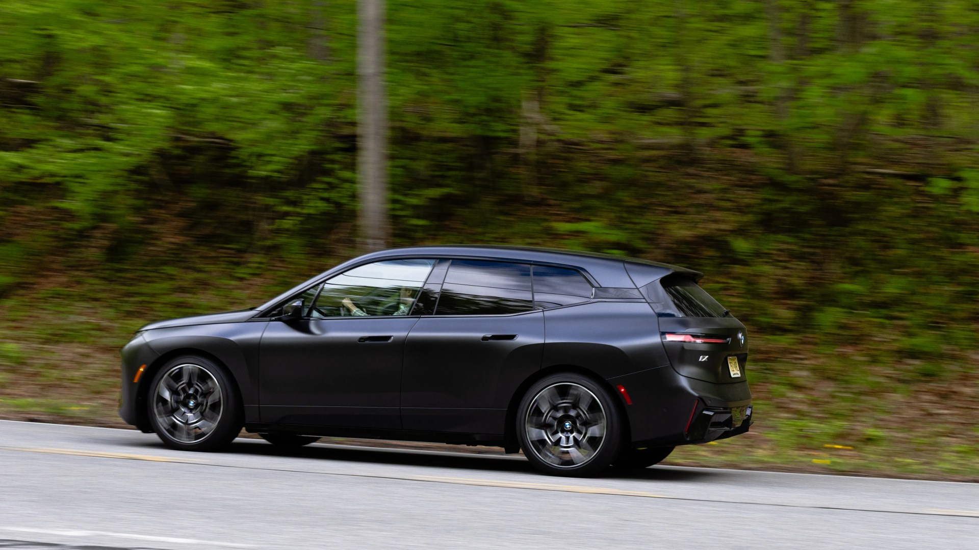 Matte black BMW iX electric SUV driving on a road with green foliage in the background.