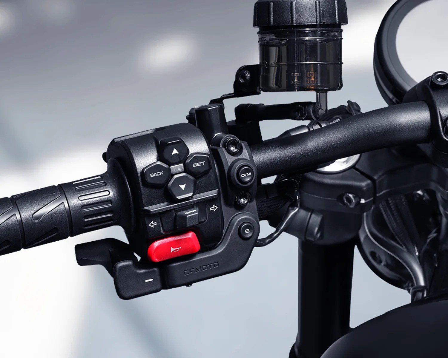 Close-up of a black motorcycle handlebar control panel with buttons, including a red horn button and a fluid reservoir above.