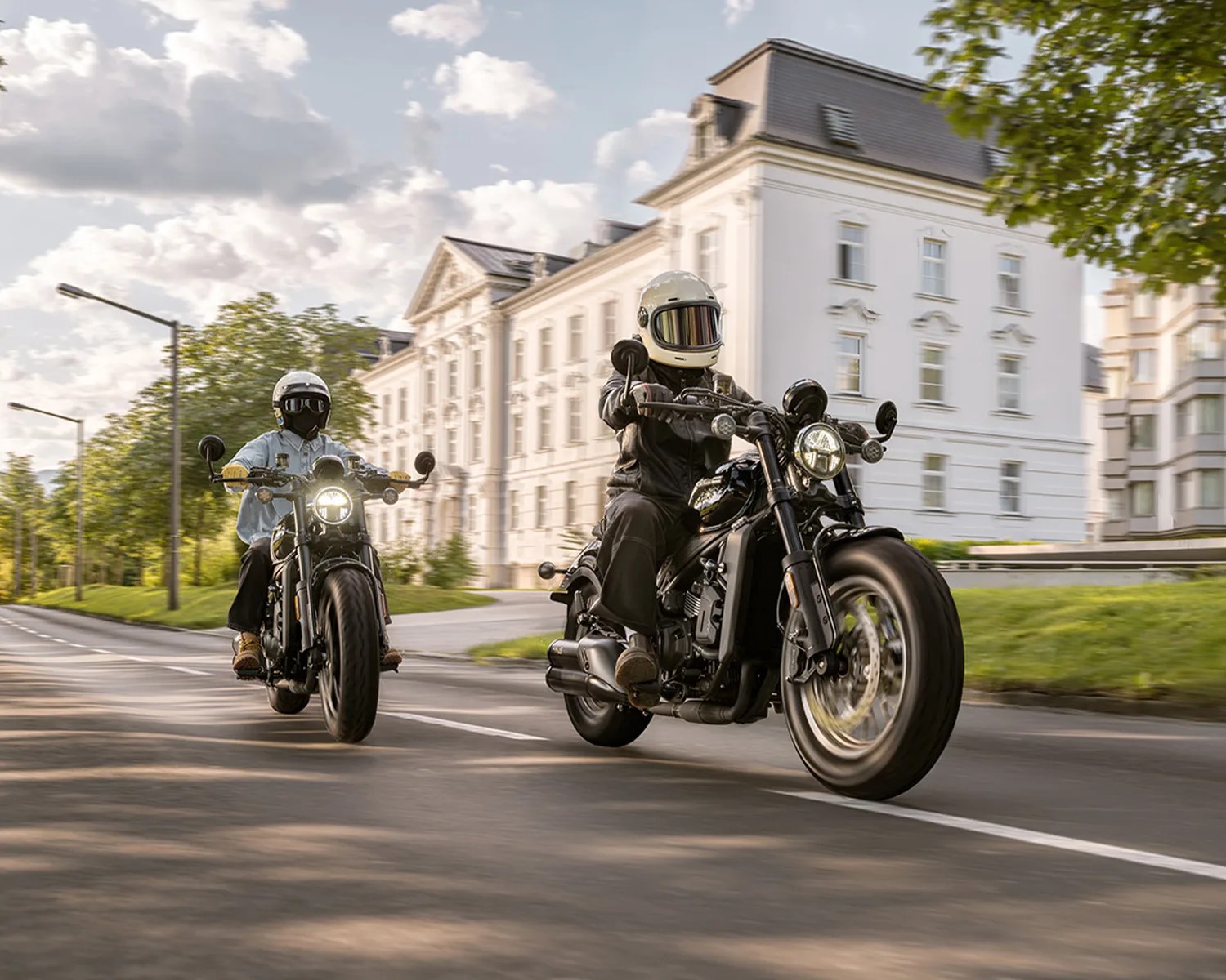 Two riders wearing helmets and jackets on black motorcycles riding on a city street with white buildings and trees.