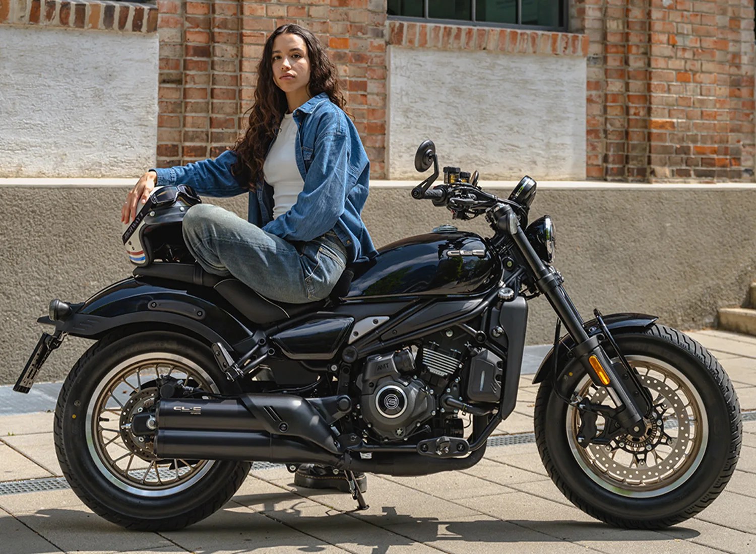 Person sitting sideways on a black motorcycle holding a black helmet with a brick wall background.