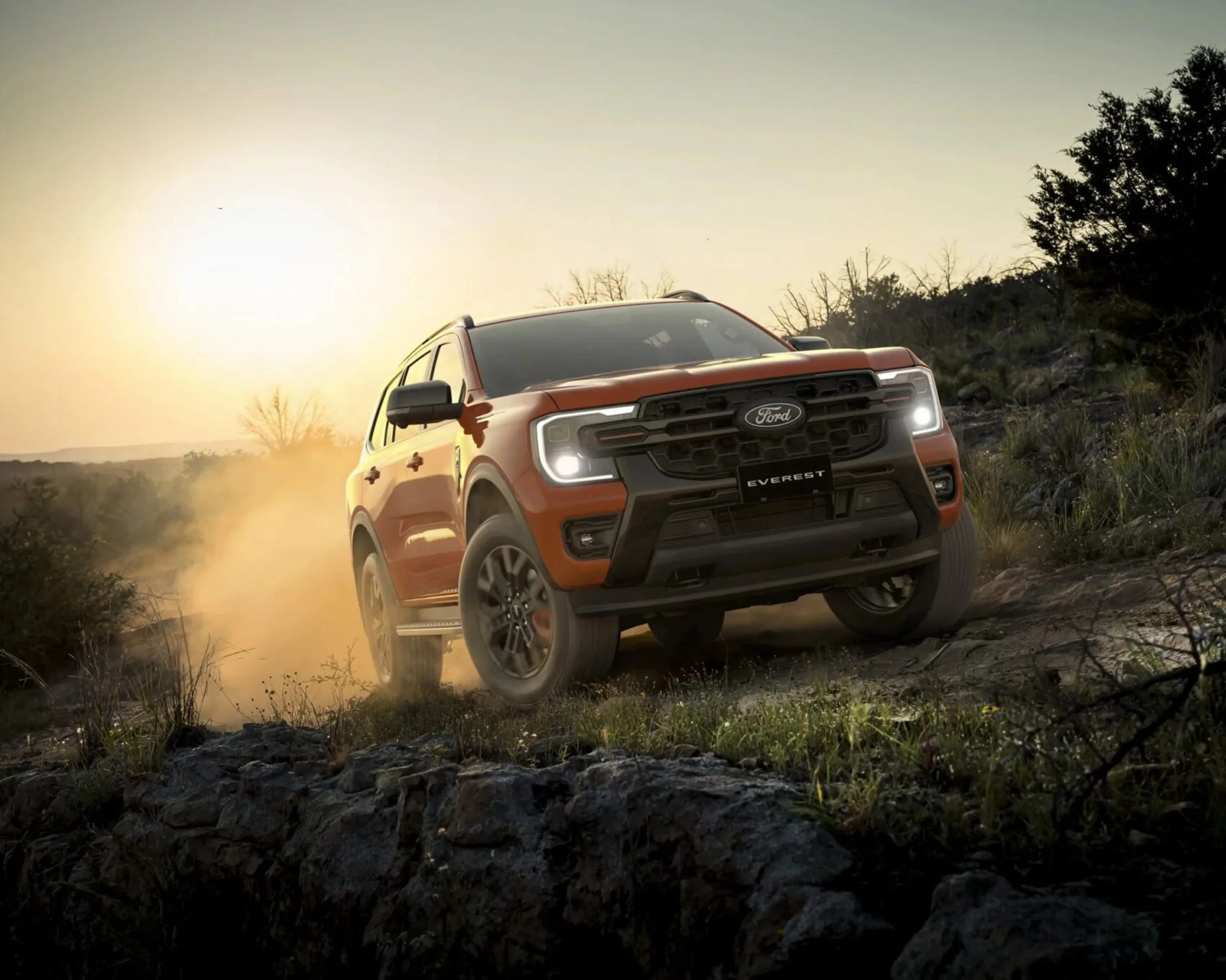 Orange Ford Everest SUV driving off-road on rocky terrain at sunset with headlights on.