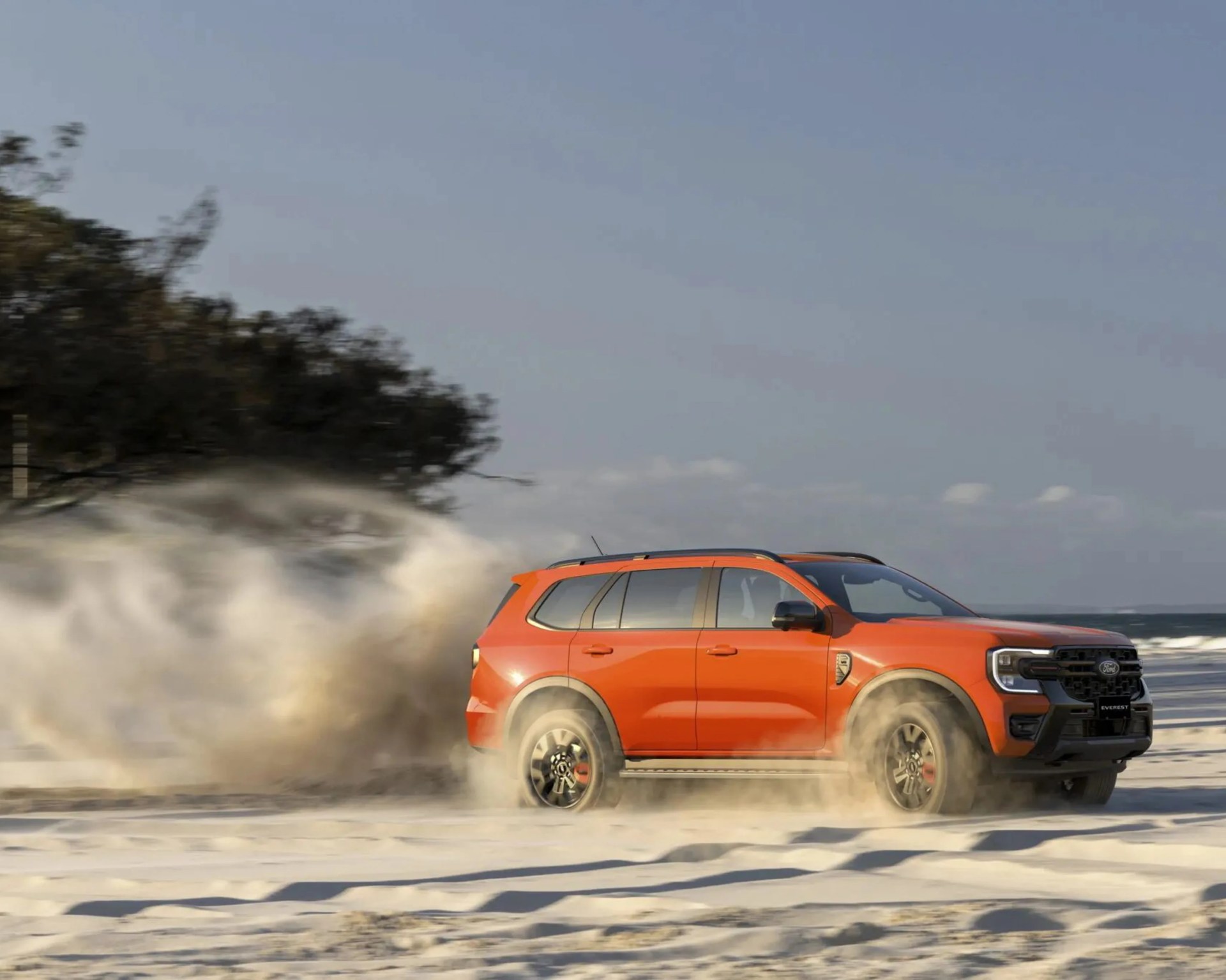 Orange Ford Everest SUV kicking up sand while driving on a beach near trees and ocean.