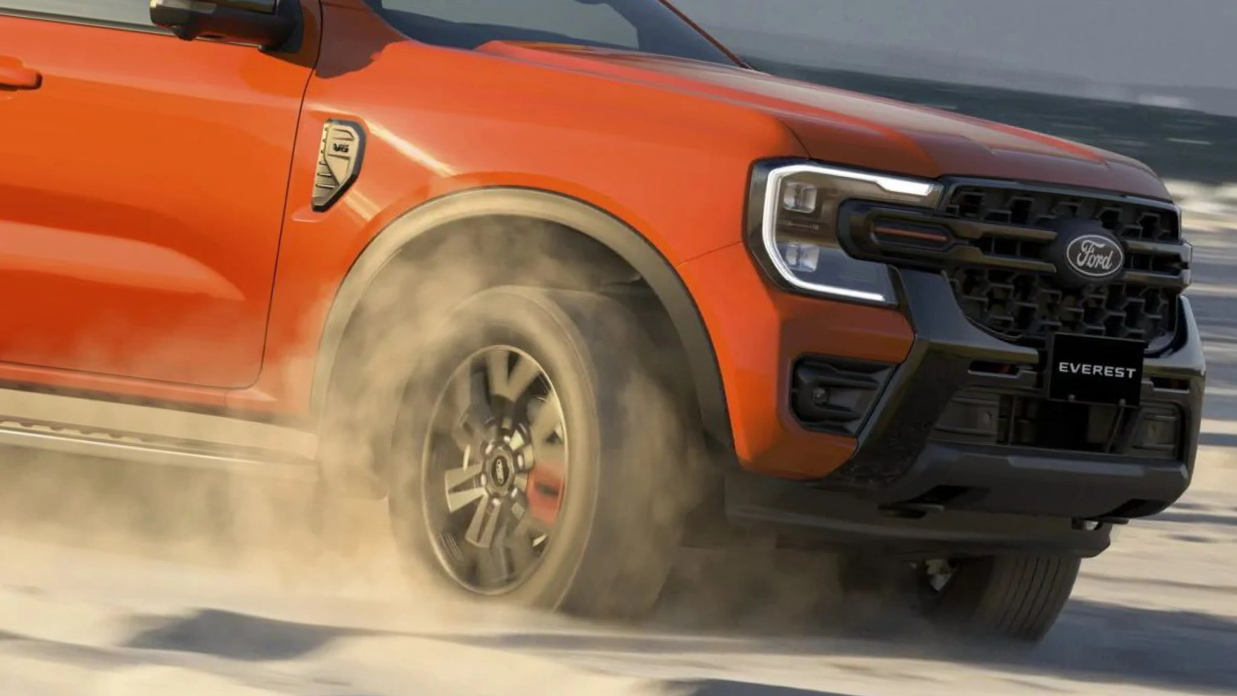 Orange Ford Everest SUV front side with spinning front wheel kicking up dust on sandy terrain.