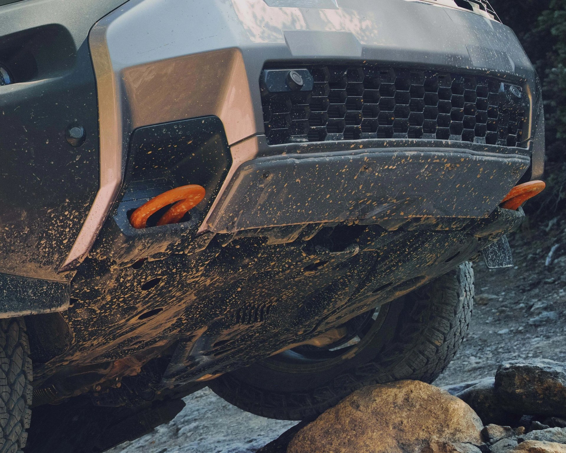 Front underside of a muddy off-road vehicle with orange tow hooks on rocky terrain.