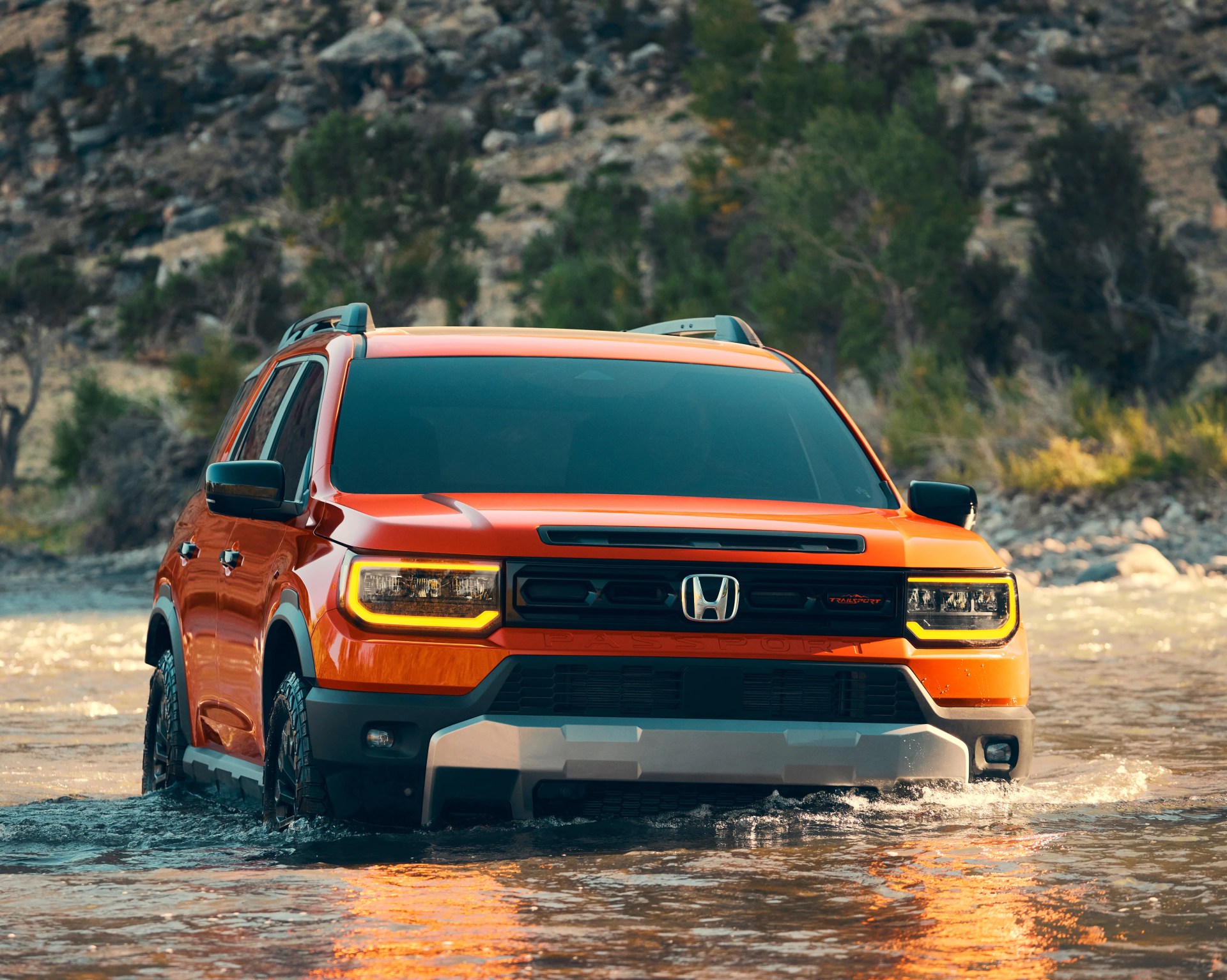 Orange Honda Passport SUV driving through shallow water with rocky hillside in background.