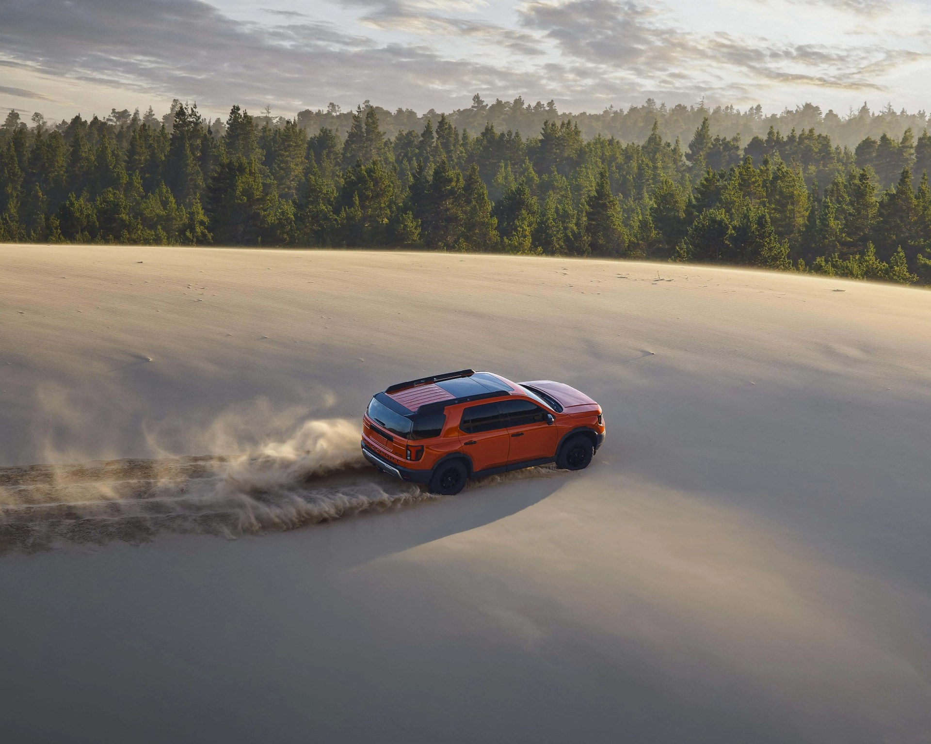 Orange SUV kicking up sand while driving on a large sand dune with a forest in the background.