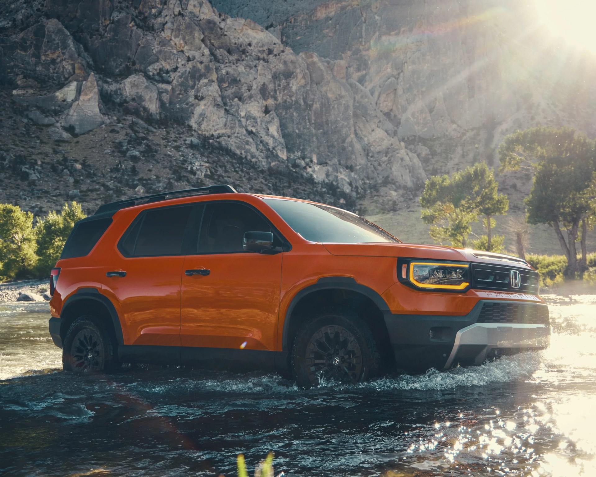 Orange Honda Passport SUV driving through shallow river with rocky mountain backdrop in sunlight.