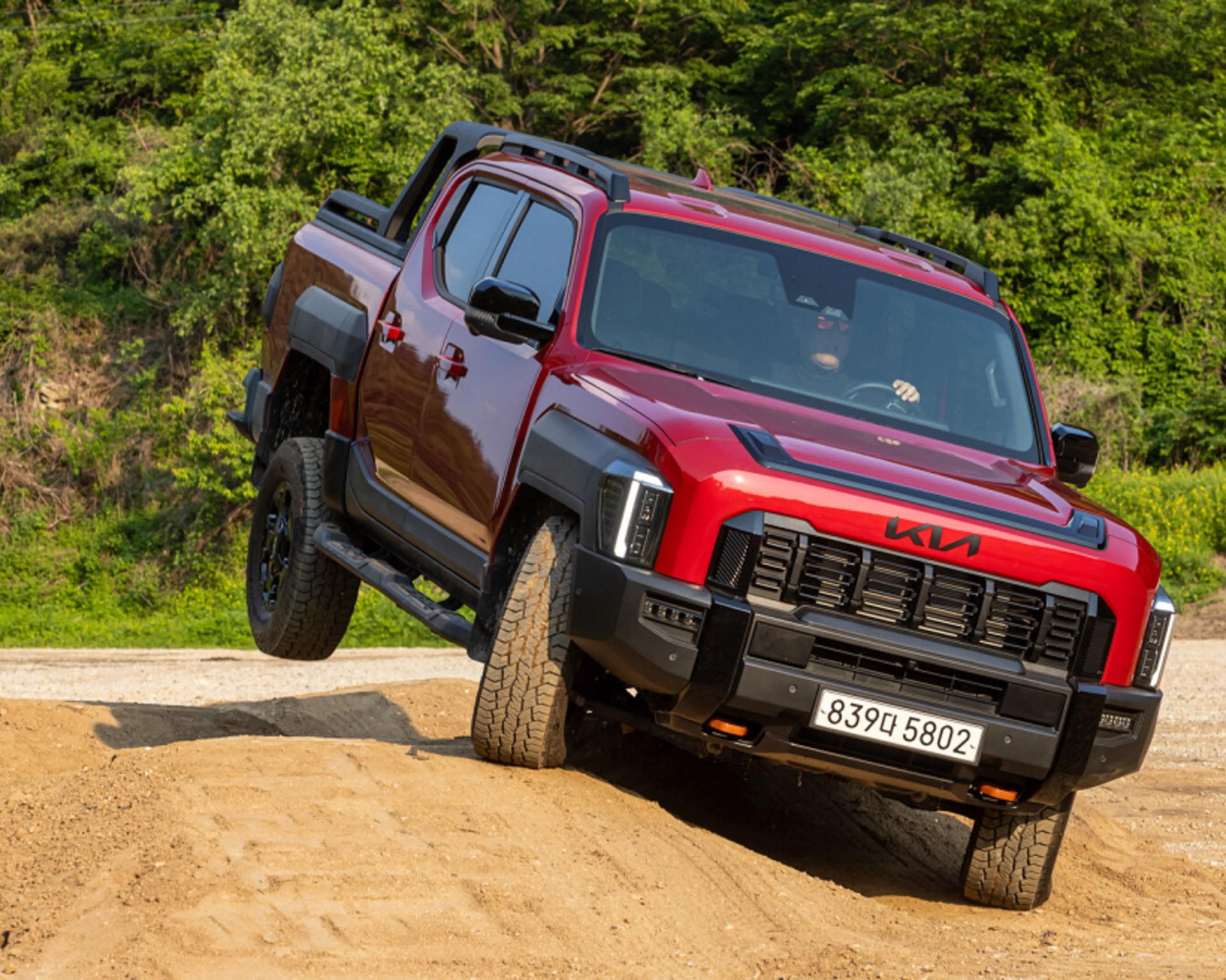 Red Kia pickup truck driving off-road on a dirt trail with one rear wheel lifted.