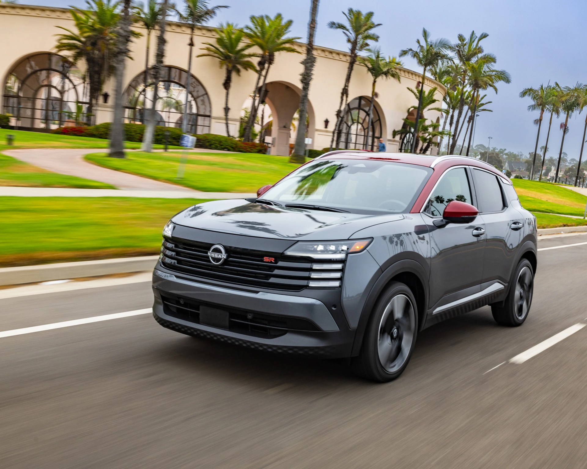 Gray Nissan SUV with red roof and side mirrors driving on a road with palm trees and a beige building in the background.