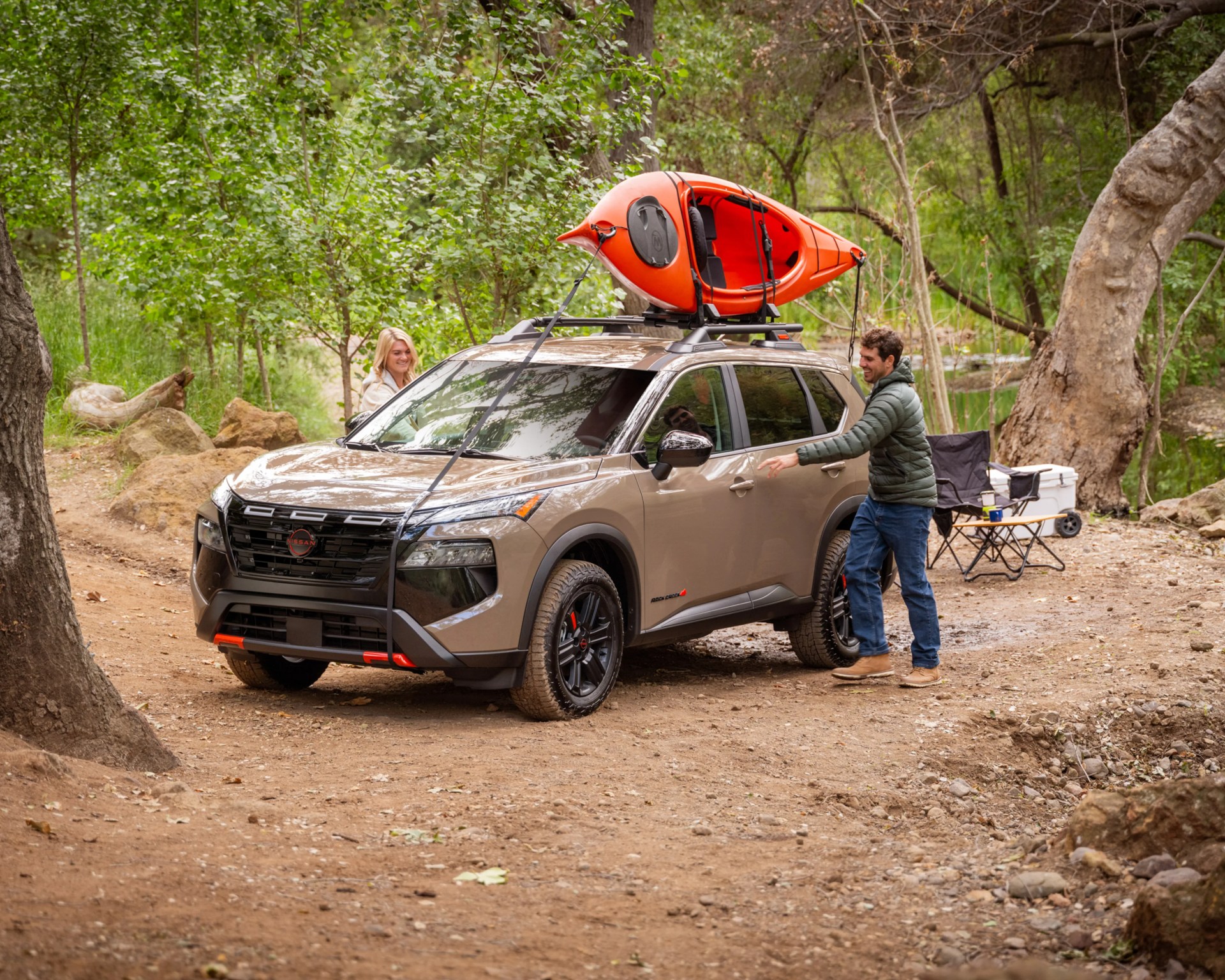 Tan Nissan SUV with black trim and roof rack carrying an orange kayak, parked on a dirt path in a forest.