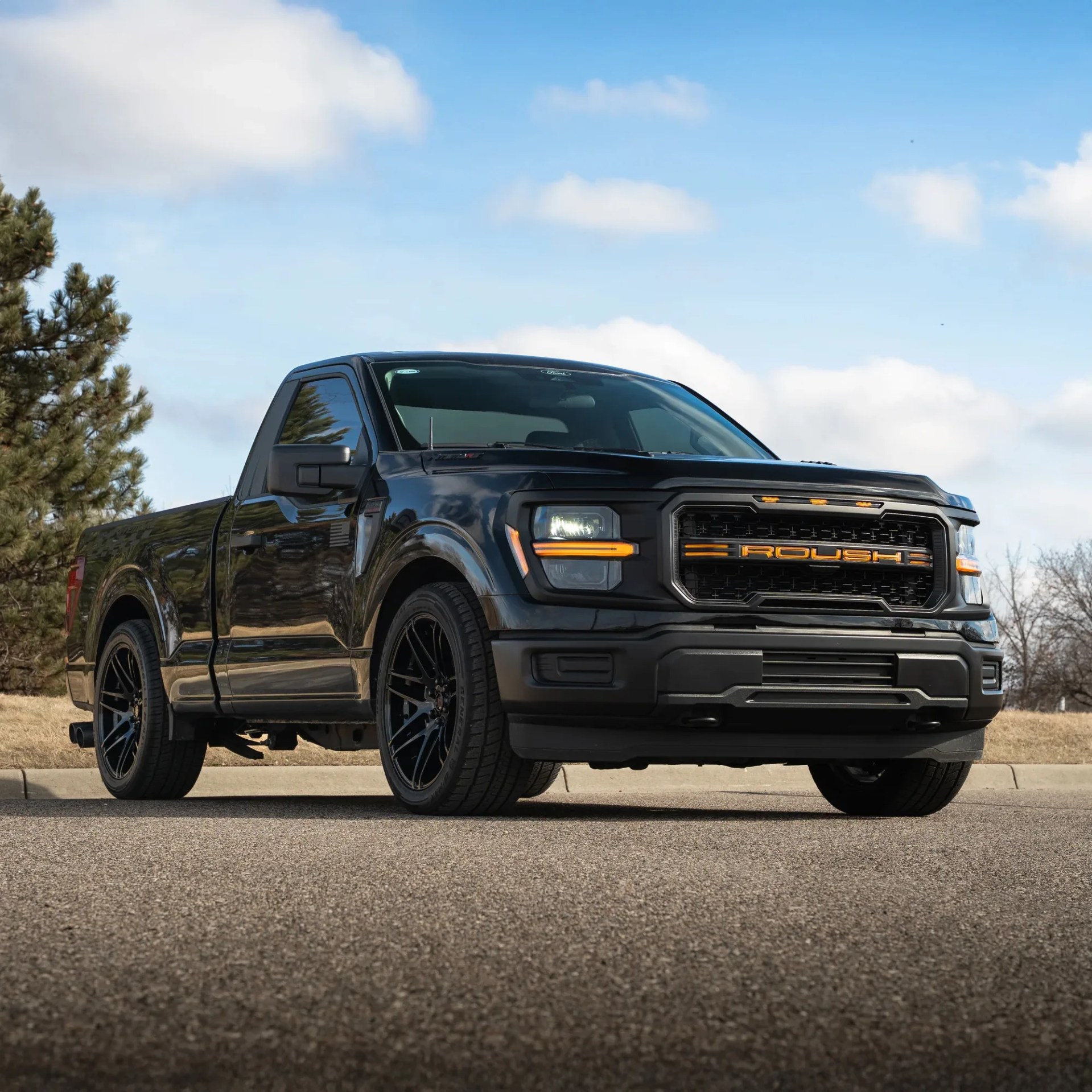 Black Roush pickup truck with orange grille lettering parked on a road under a blue sky.