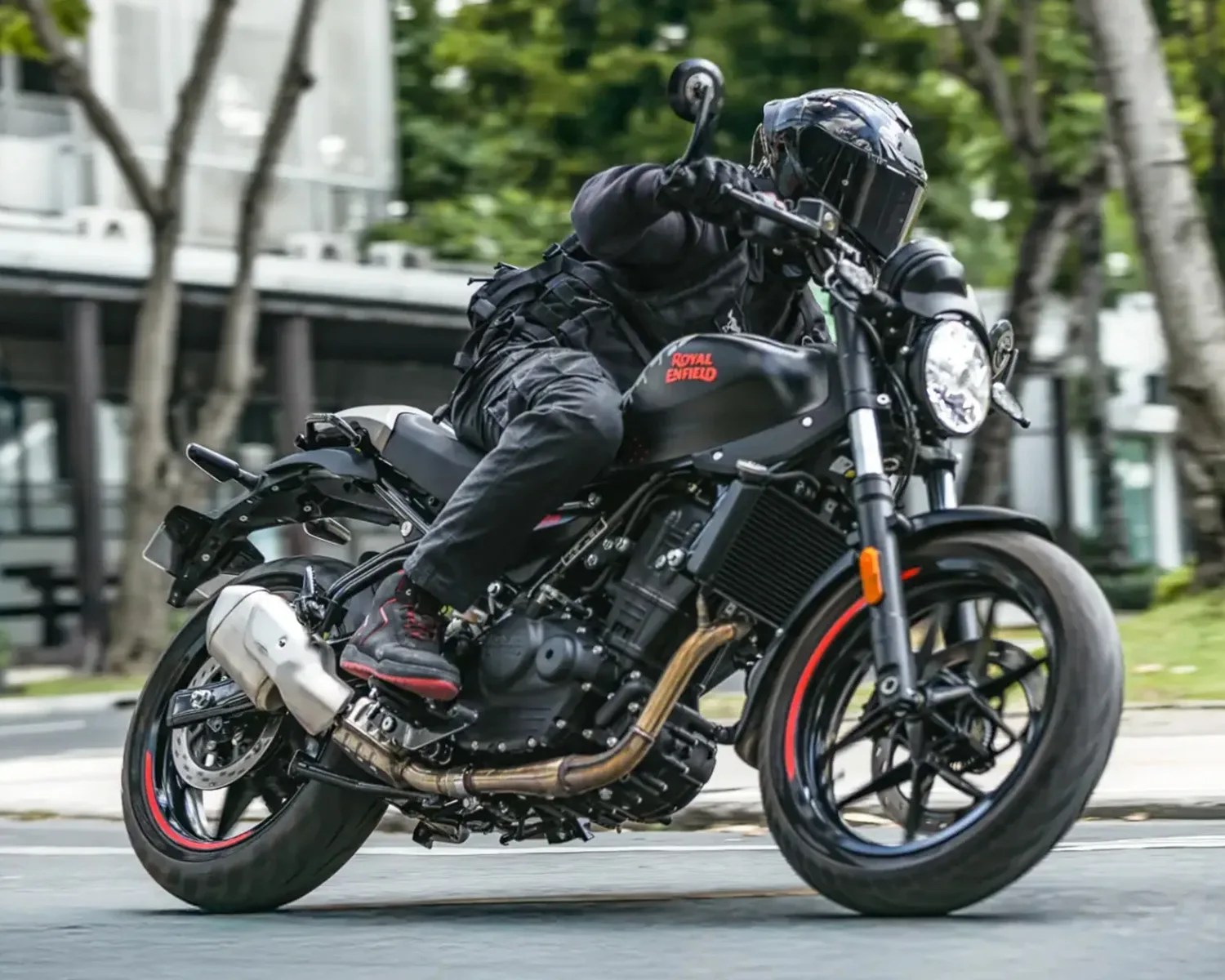 Person wearing black gear riding a black Royal Enfield motorcycle with red accents on a city street.