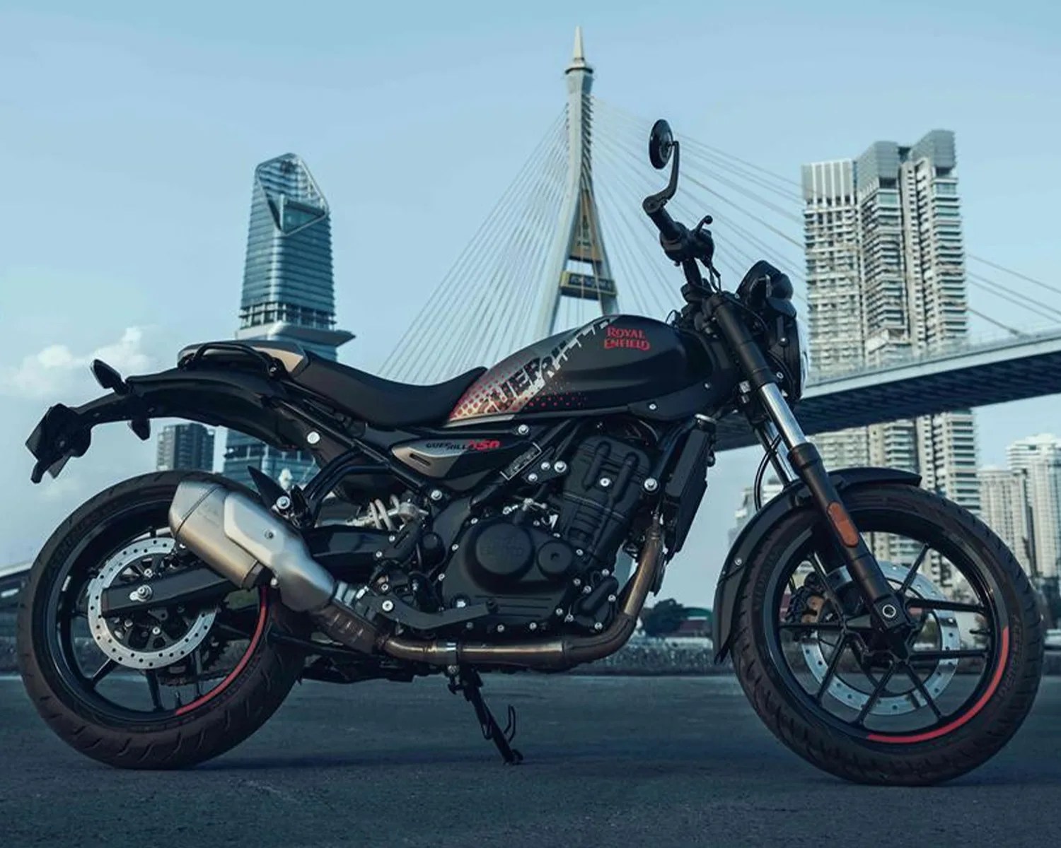 Black Royal Enfield motorcycle with red accents parked on a city street with a cable-stayed bridge and skyscrapers in the background.