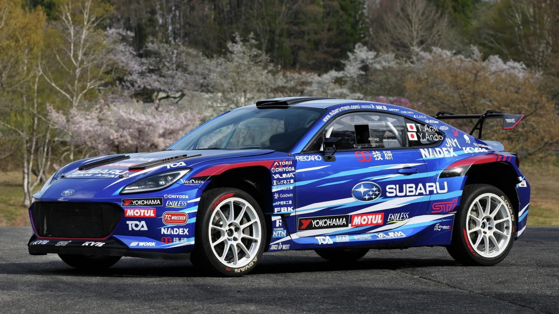 Blue Subaru rally car with white wheels and multiple sponsor logos parked on asphalt with trees in the background.