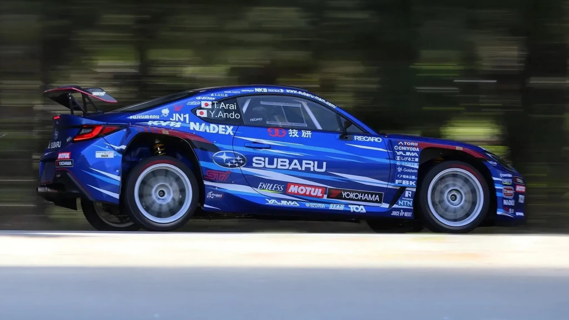 Blue Subaru race car with sponsor logos and a large rear spoiler in motion on a road.