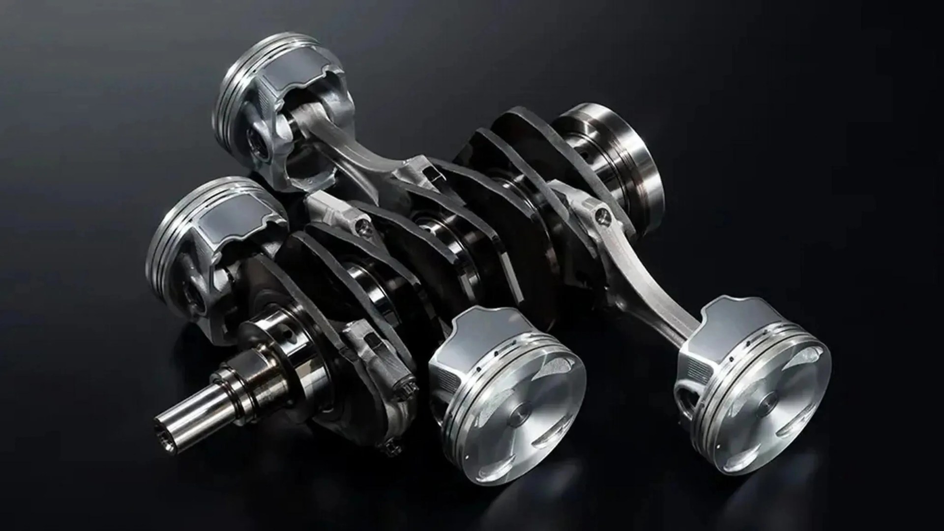 Polished metal crankshaft with four attached pistons on a dark reflective surface.