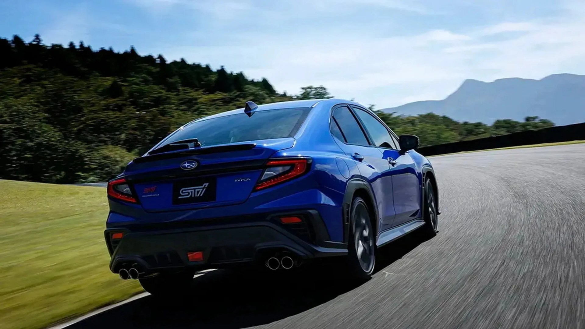 Blue Subaru WRX STI sedan driving on a curved road with green trees and mountains in the background.