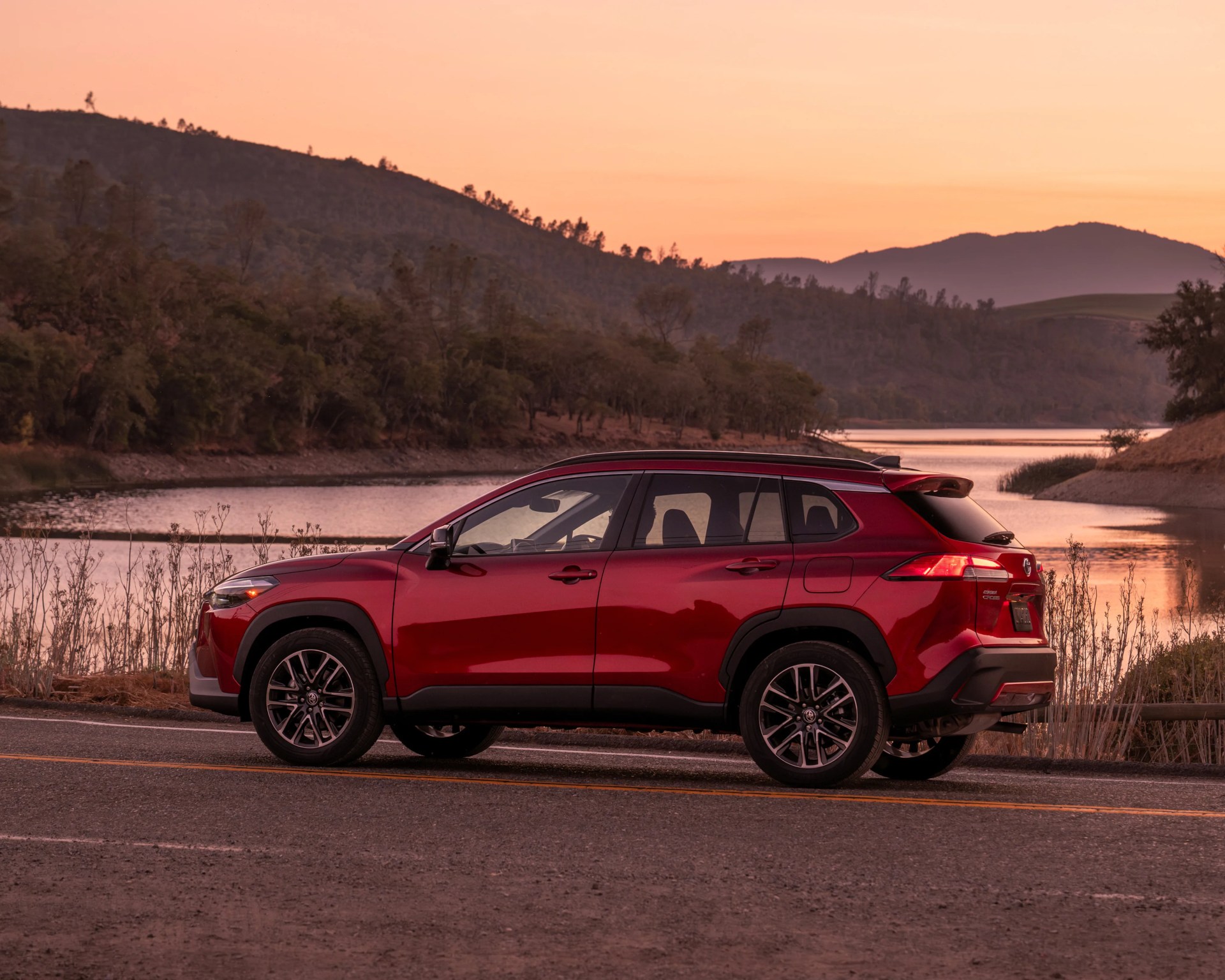 Red Toyota Corolla Cross SUV parked on a road near a lake at sunset with hills in the background.
