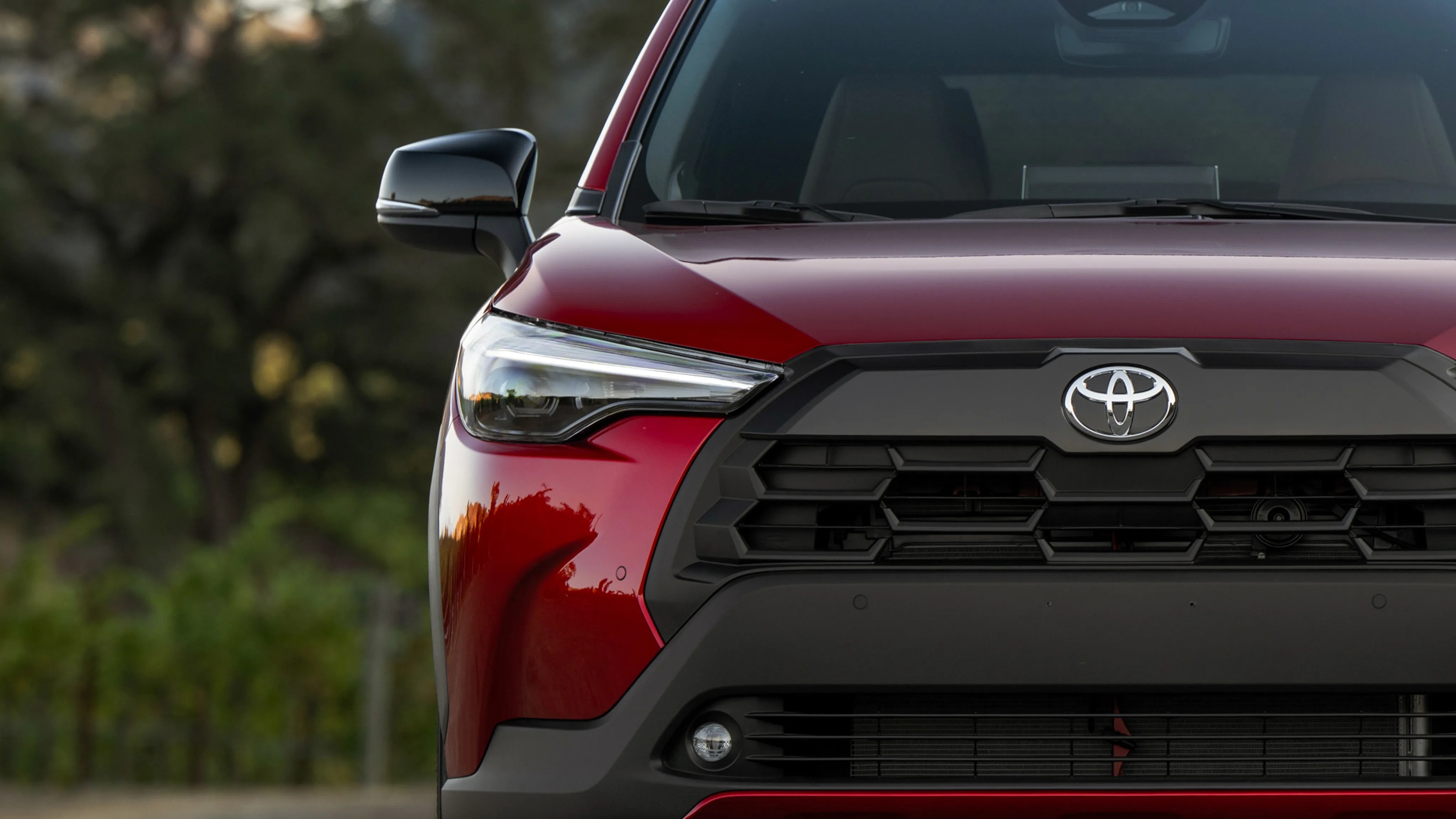 Front view of a red Toyota vehicle showing the left headlight, side mirror, and black grille with the Toyota emblem.
