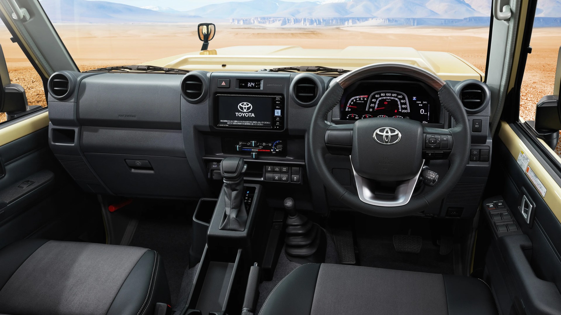 Interior view of a Toyota Land Cruiser dashboard with black steering wheel, digital display, and gear shifter in a desert setting.