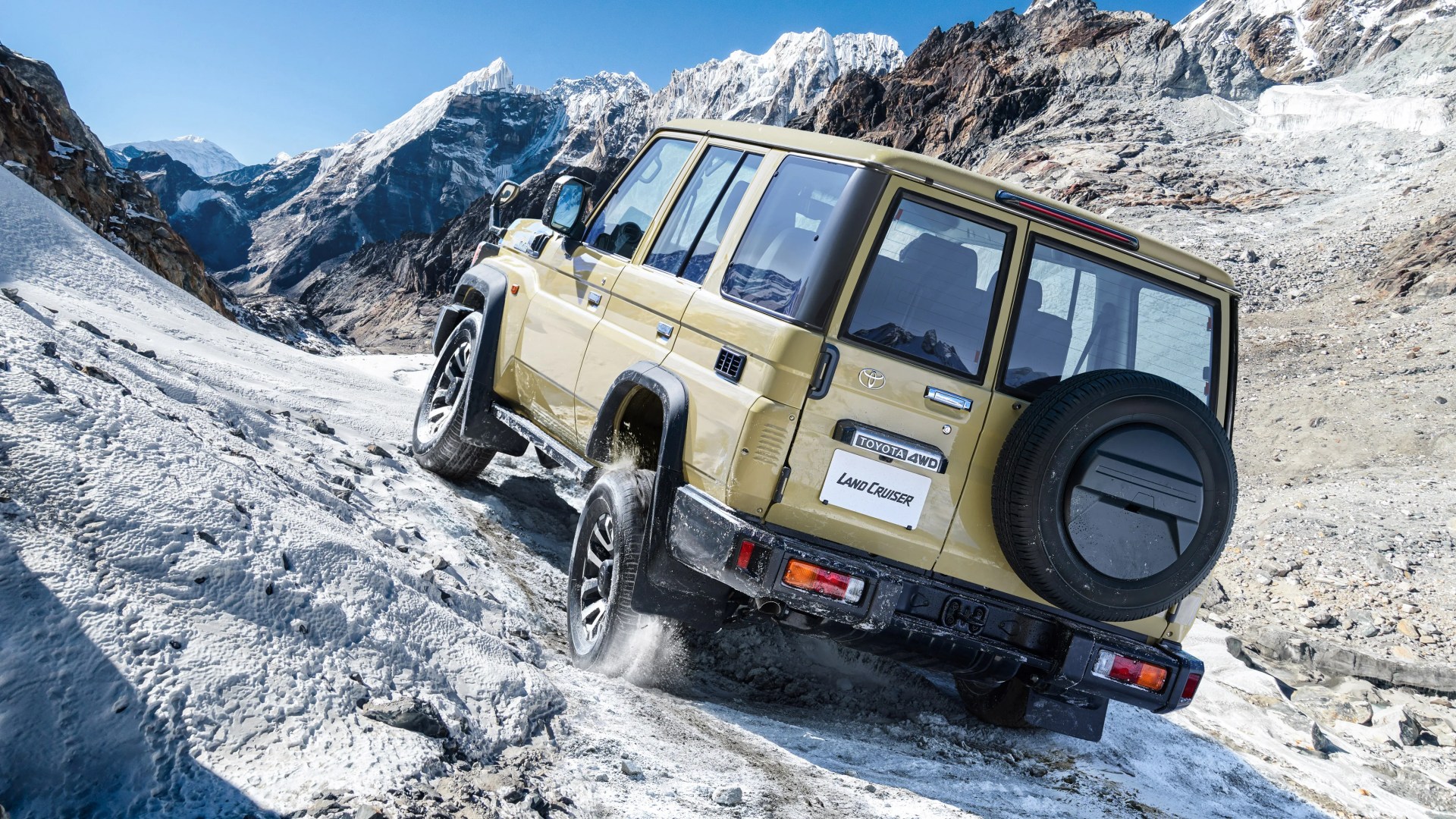 Beige Toyota Land Cruiser 4WD driving uphill on a snowy mountain trail with rocky peaks in the background.