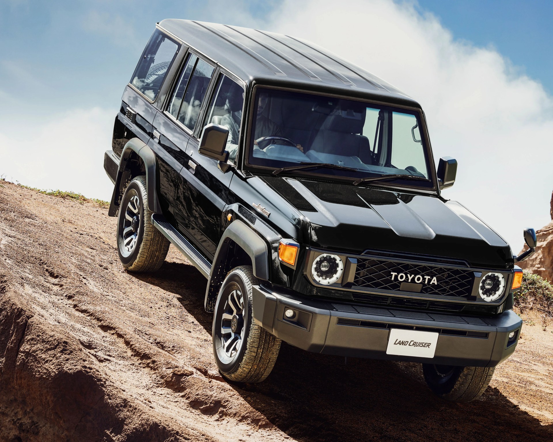 Black Toyota Land Cruiser SUV driving on a steep dirt hill under a blue sky.