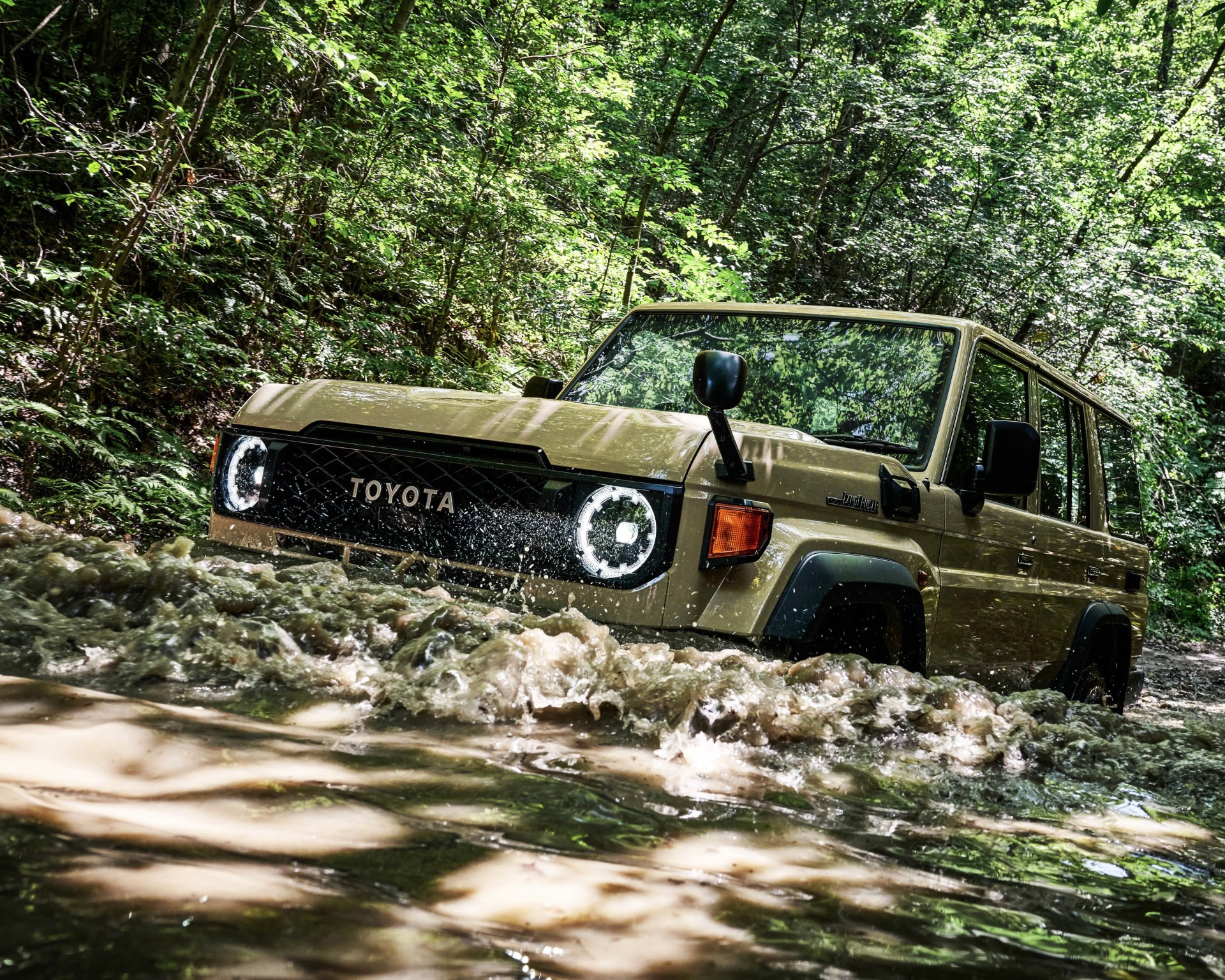 Beige Toyota Land Cruiser driving through a shallow, muddy river surrounded by dense green forest.