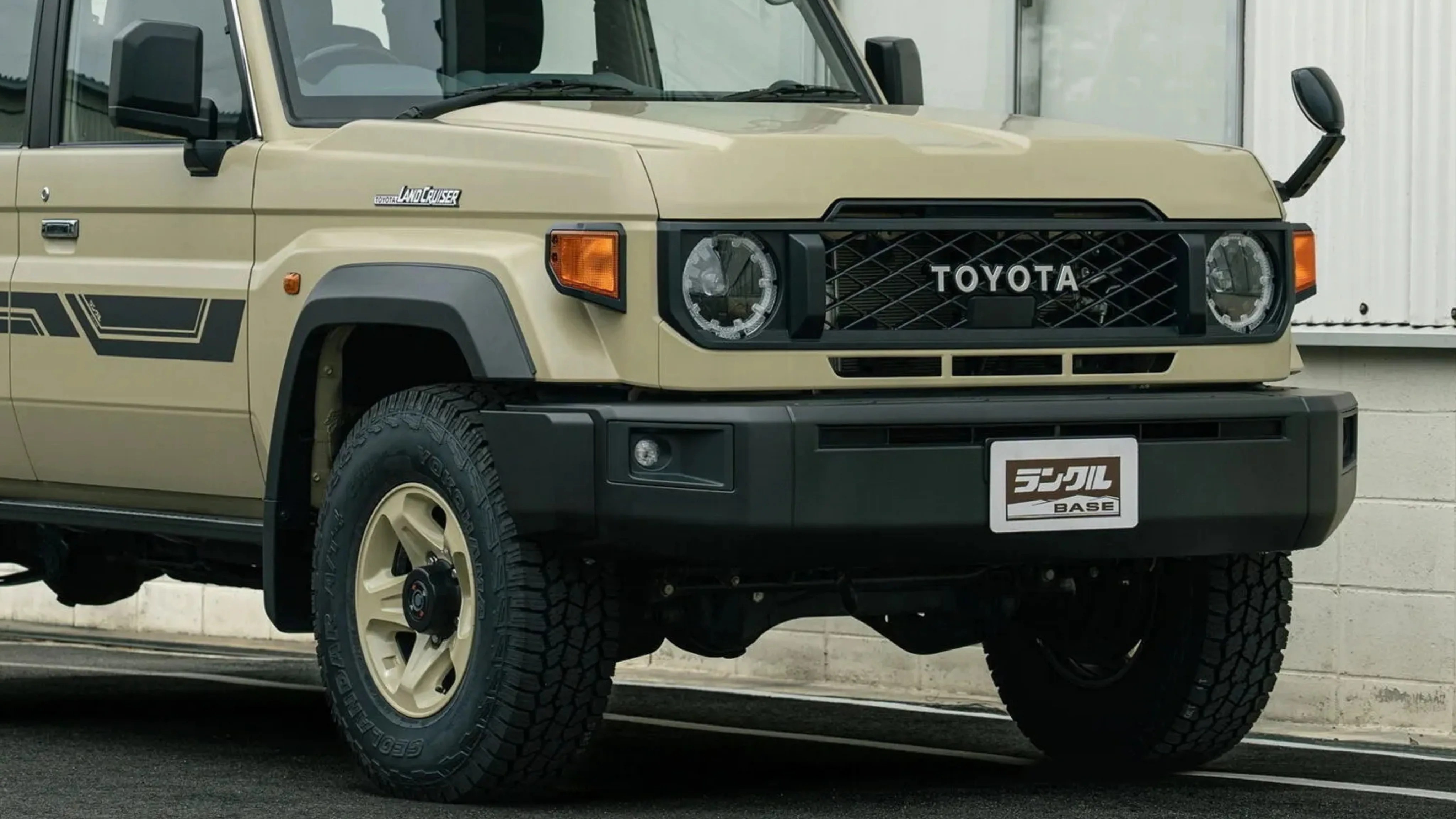 Beige Toyota Land Cruiser with black grille, round headlights, and off-road tires parked by a white wall.