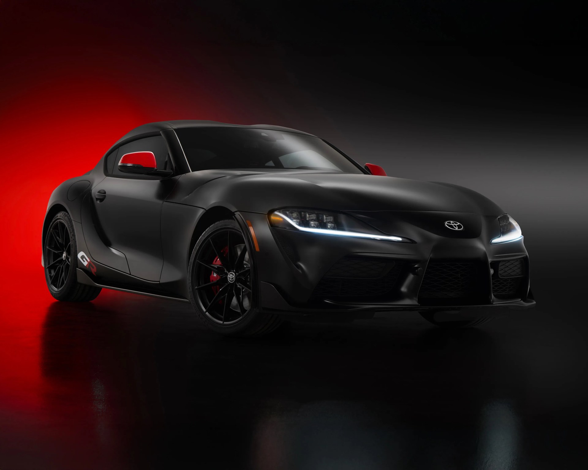 Matte black Toyota GR Supra sports car with red side mirrors and brake calipers, illuminated headlights, on a dark red and black background.