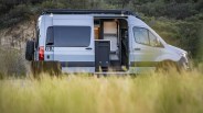 White camper van with sliding door open showing interior kitchen and seating, parked in grassy area with trees in background.