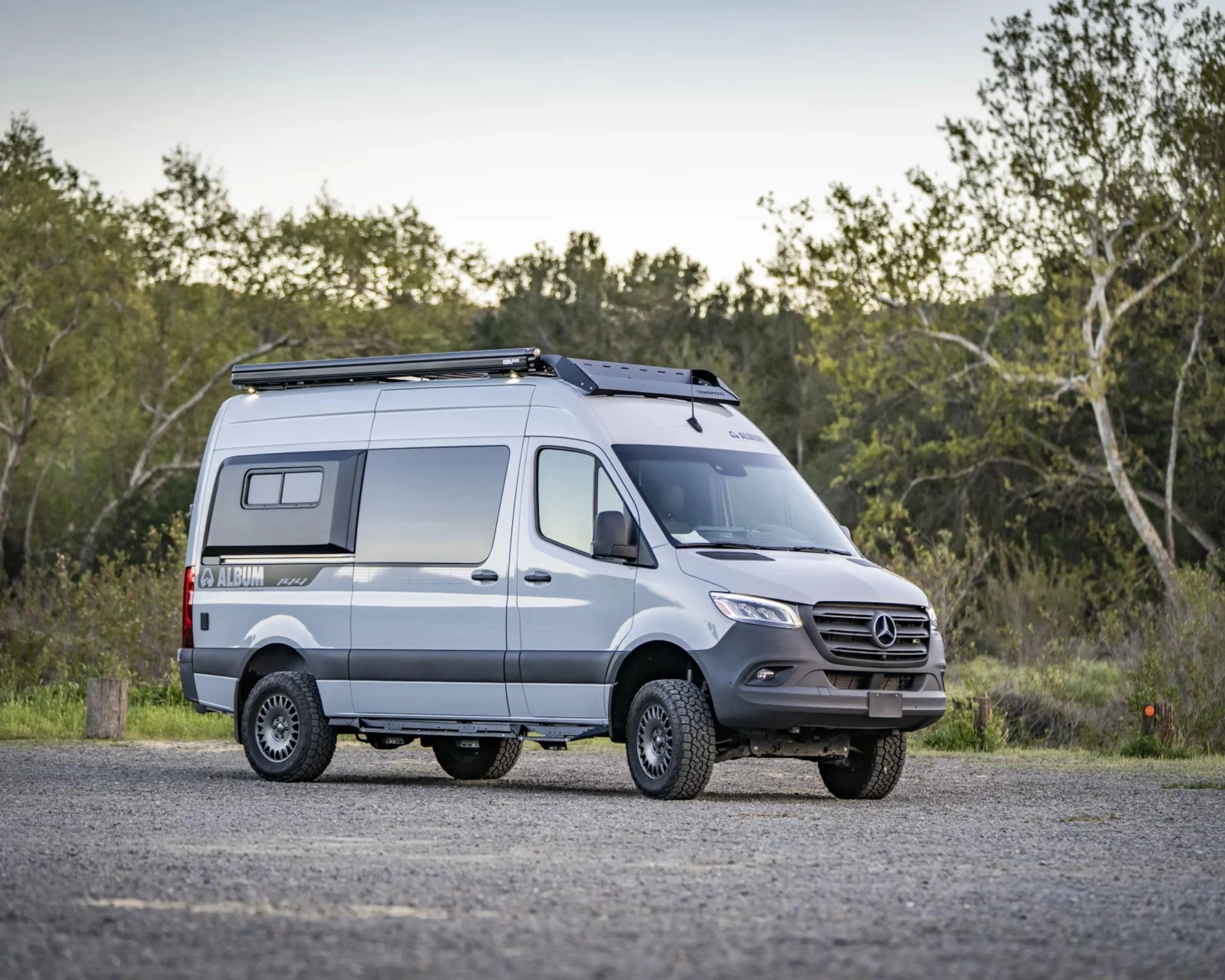 Silver Mercedes-Benz camper van with off-road tires parked on gravel near trees.