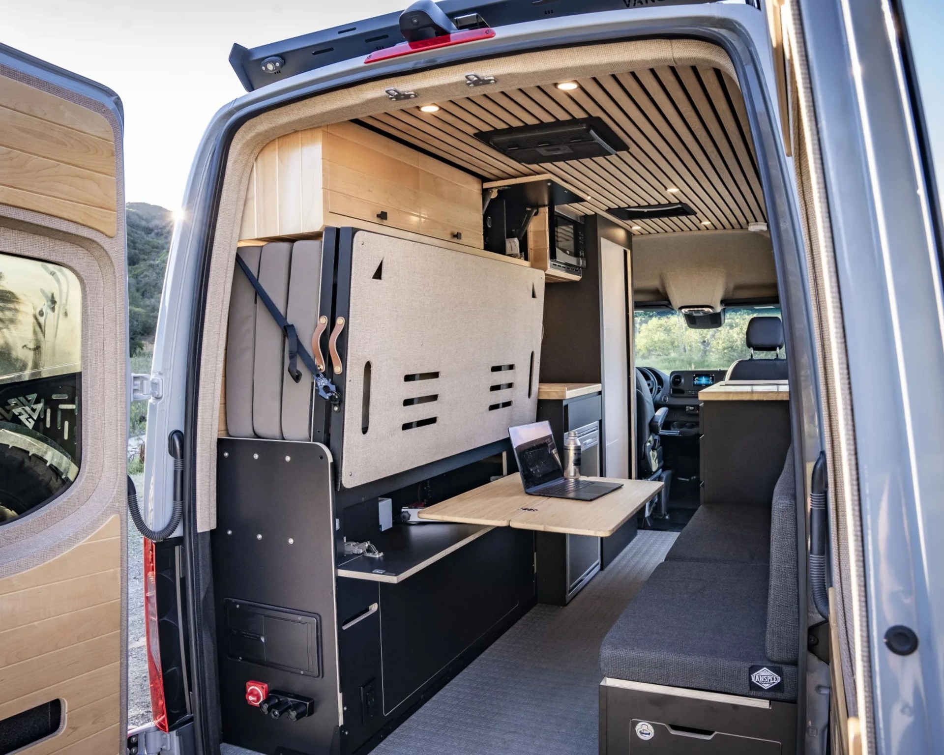Interior view of a camper van with wooden ceiling, foldable table with laptop, cushioned bench, and storage cabinets.