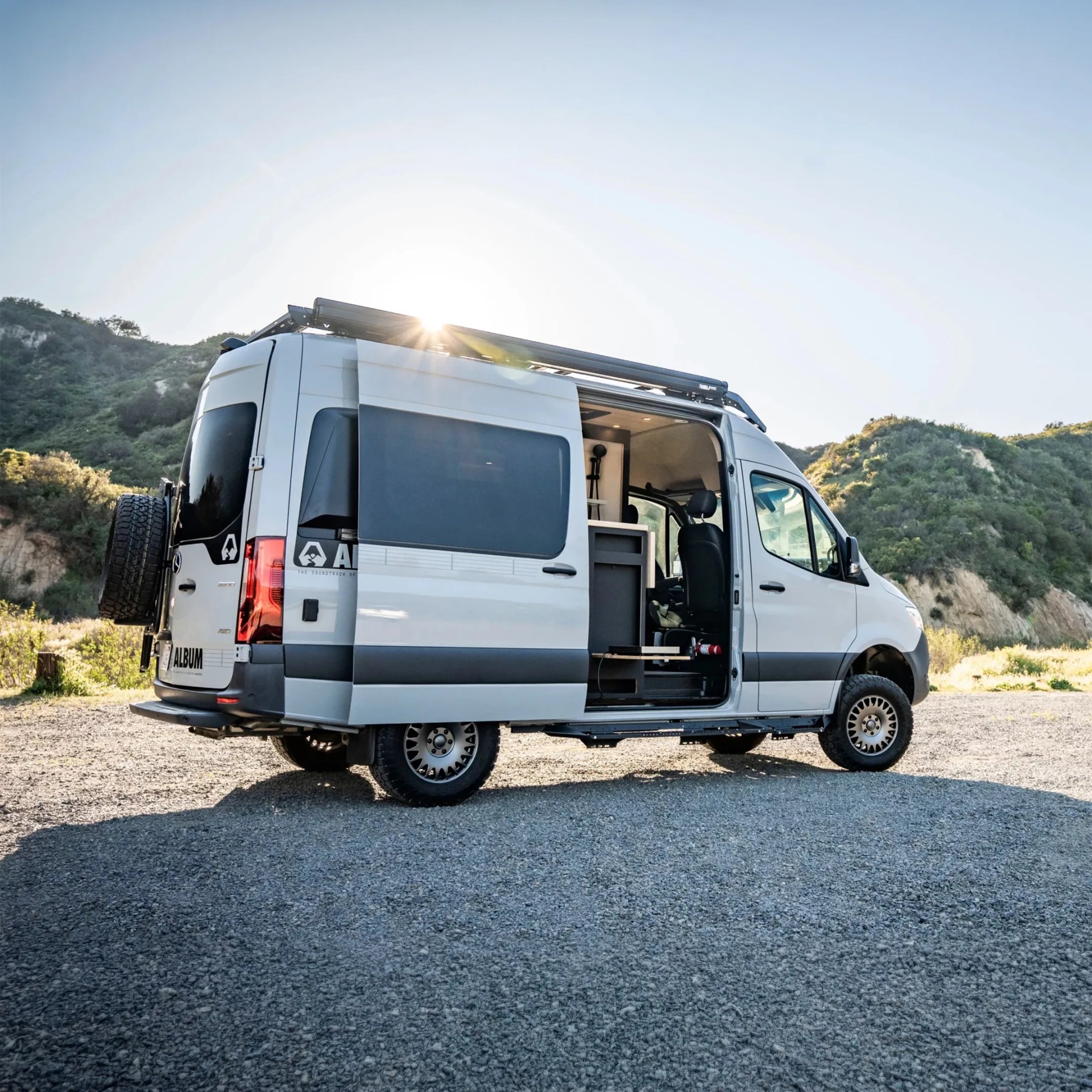 White camper van with open sliding door showing interior kitchen and seating, parked on gravel near green hills.