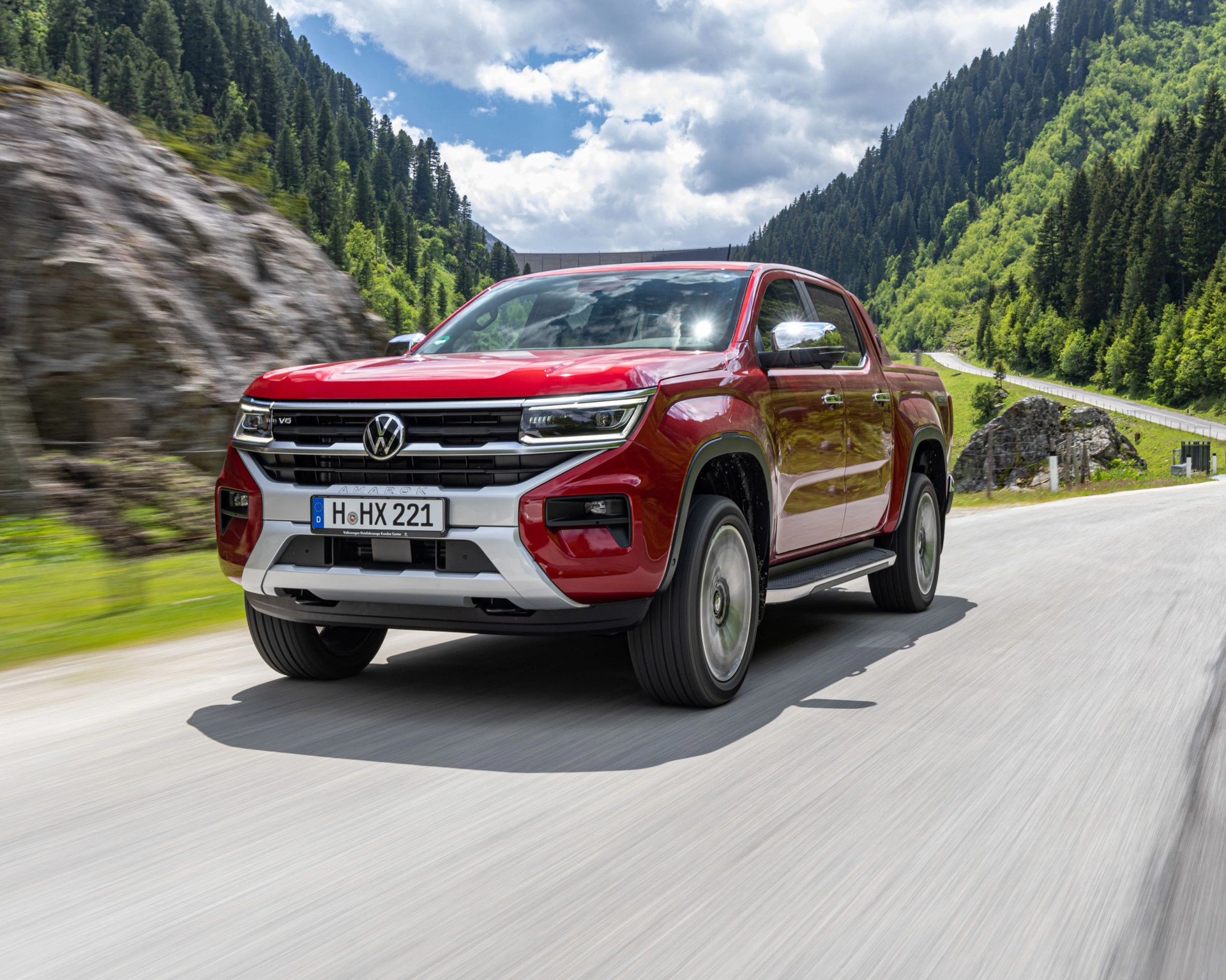 Red Volkswagen Amarok pickup truck driving on a mountain road with green trees and rocky hillsides.