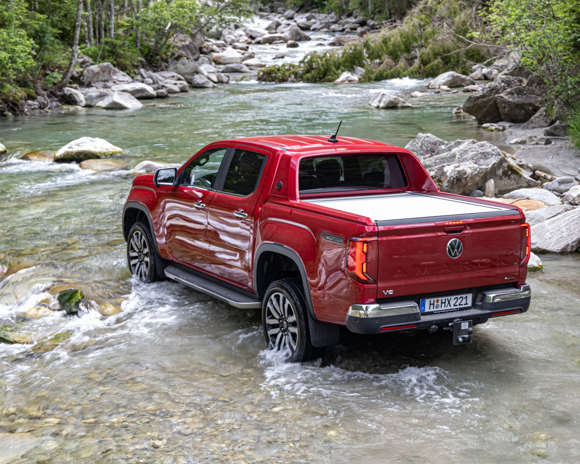 Red Volkswagen Amarok V6 pickup truck driving through a shallow rocky river surrounded by trees.