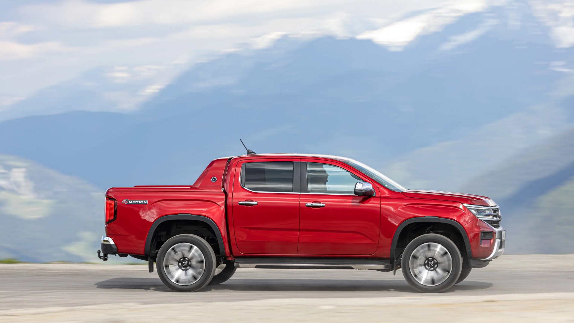 Red Volkswagen Amarok pickup truck driving on a mountain road with blurred background.