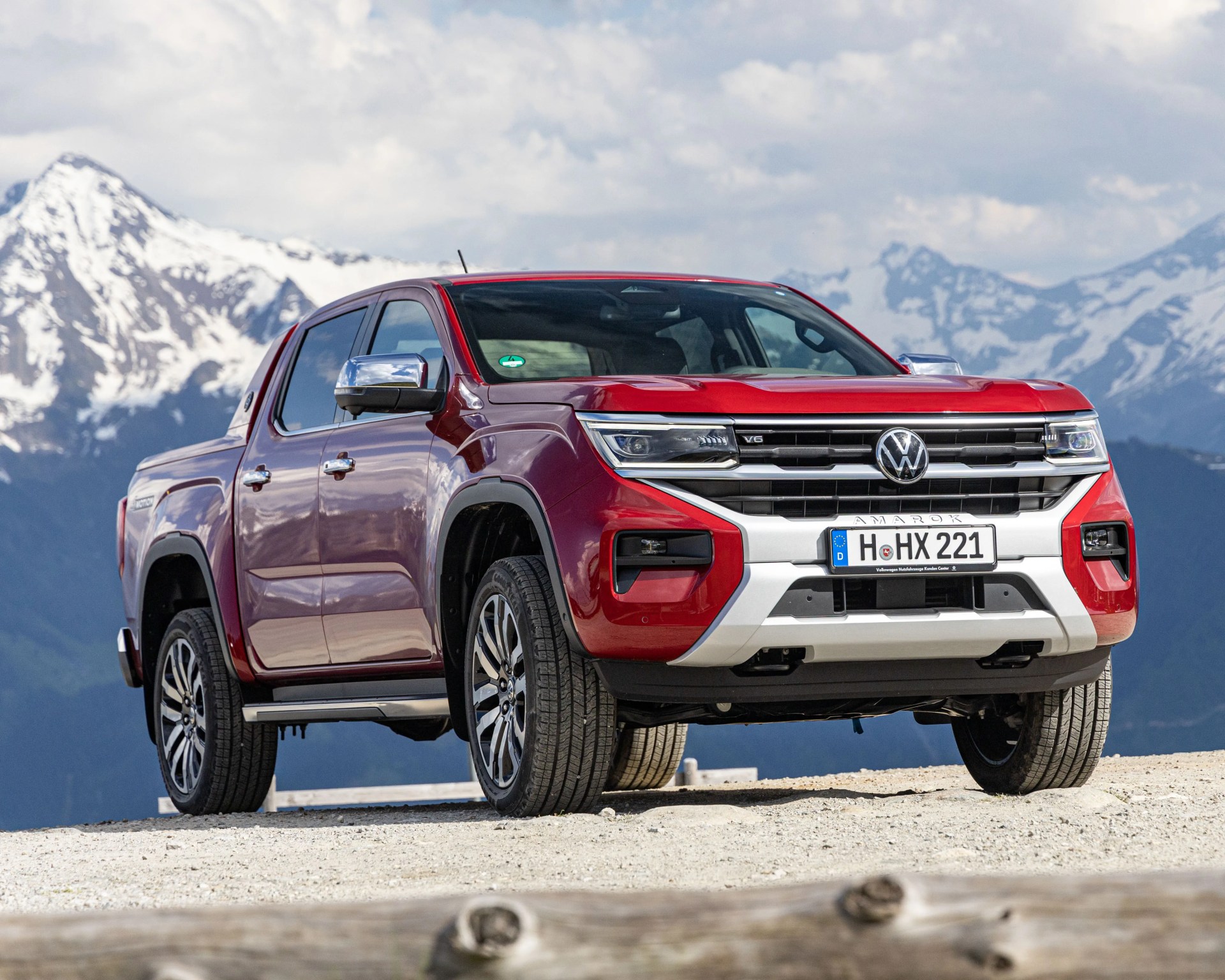 Red Volkswagen Amarok V6 pickup truck parked on a gravel surface with snow-capped mountains in the background.