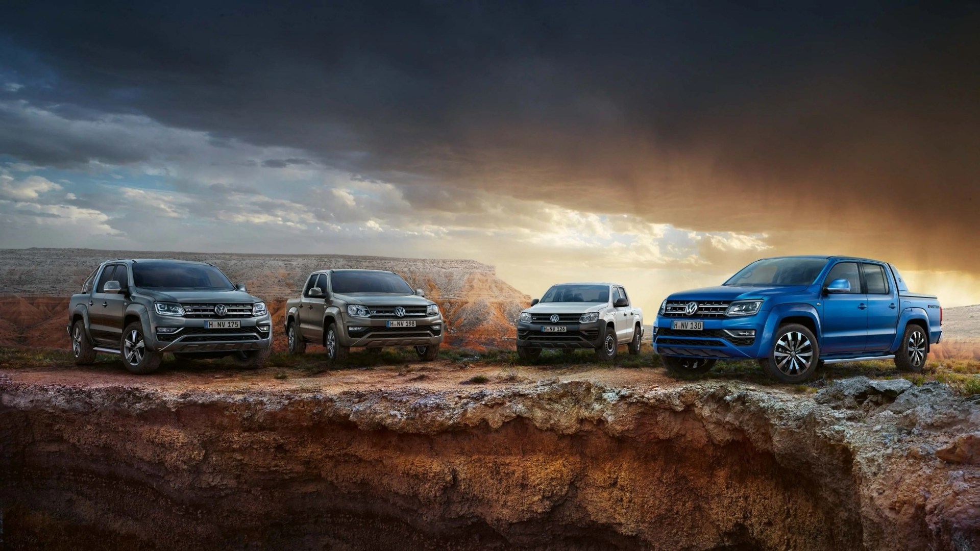 Four Volkswagen pickup trucks in gray, beige, white, and blue parked on rocky terrain under a dramatic cloudy sky.