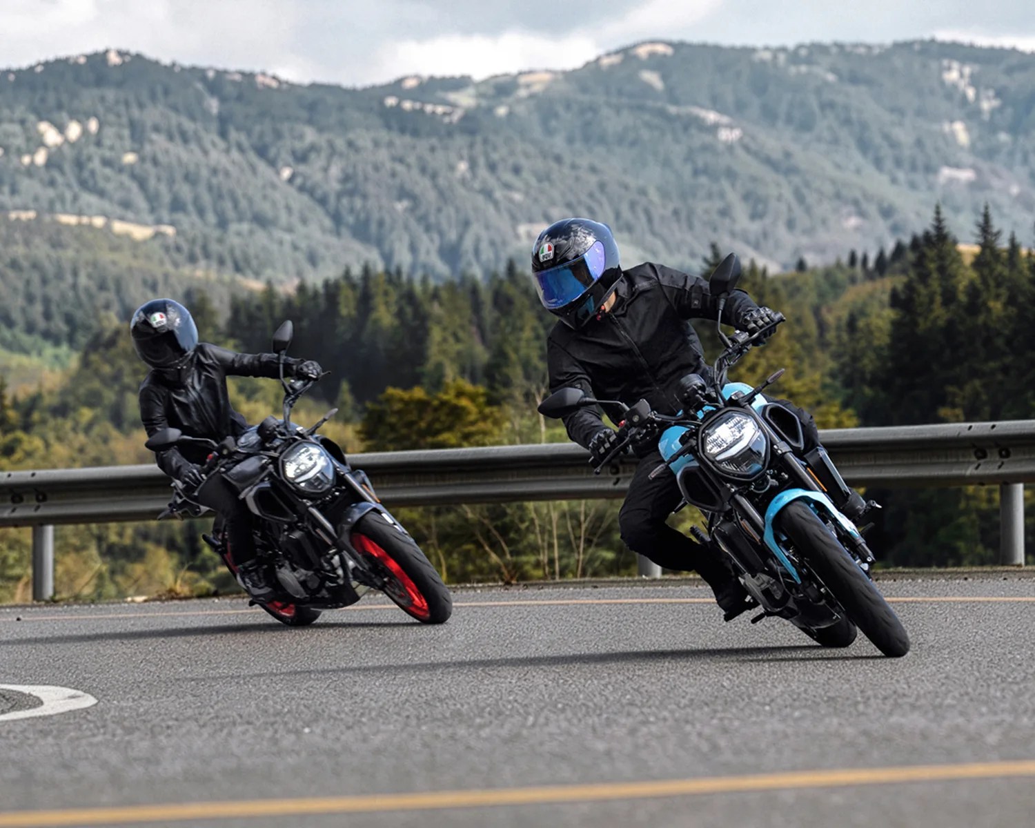 Two motorcyclists wearing black gear and helmets leaning into a curve on a mountain road, one bike with blue accents and the other with red wheels.