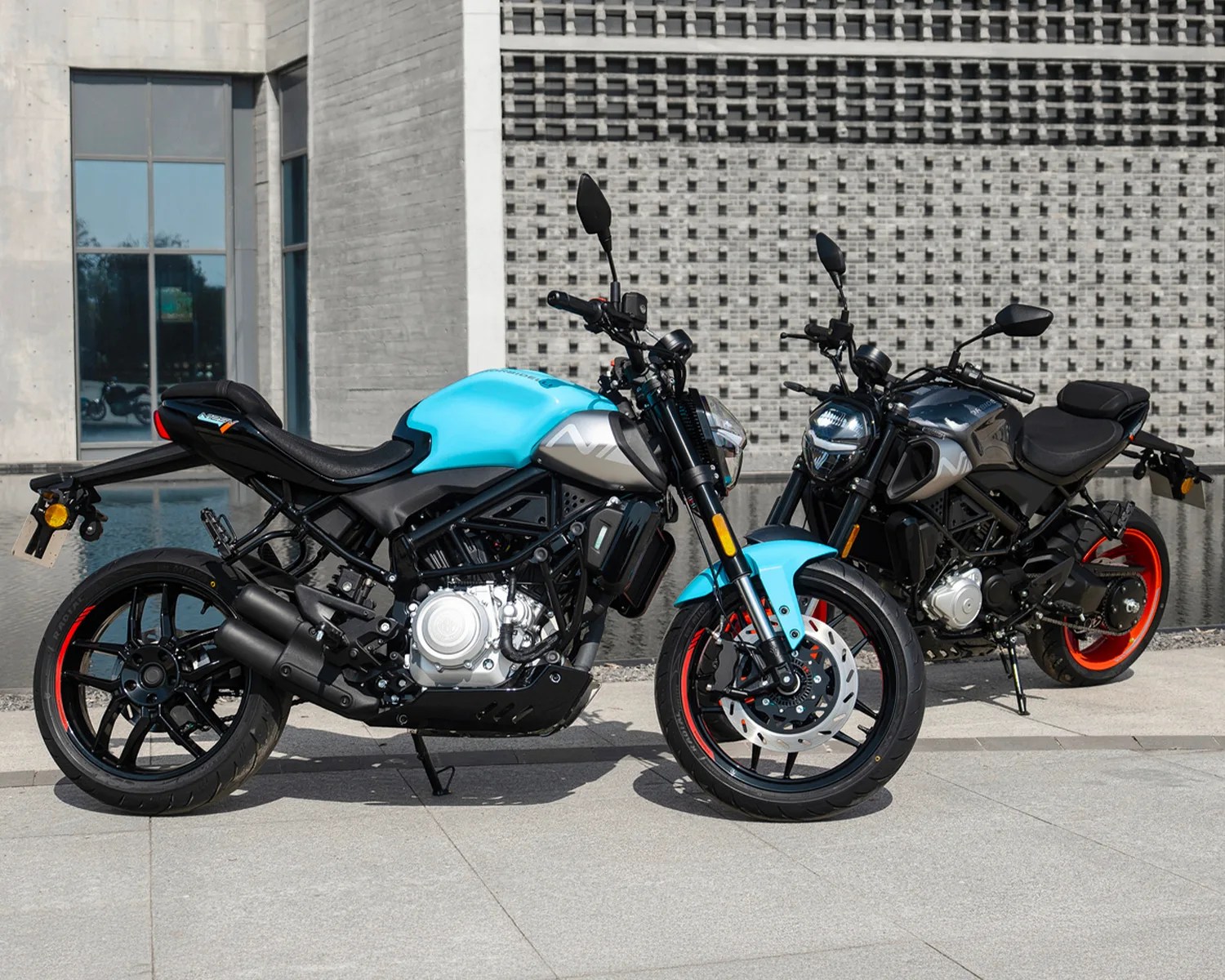 Two modern motorcycles, one with a turquoise fuel tank and the other with a dark gray fuel tank, both with black frames and red-rimmed wheels, parked on a concrete surface near a building with a textured wall.