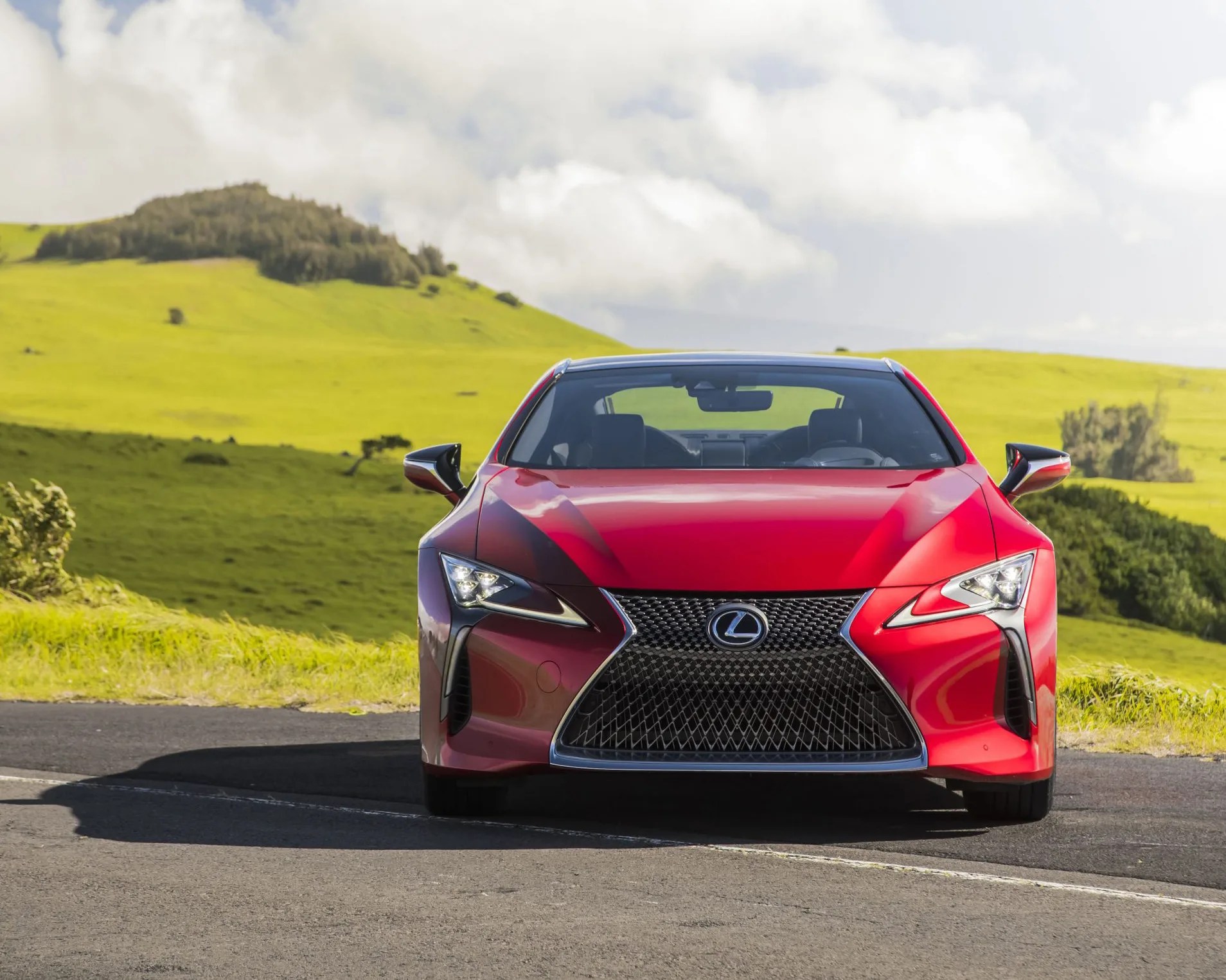 Front view of a red Lexus car parked on a road with green hills and cloudy sky in the background.