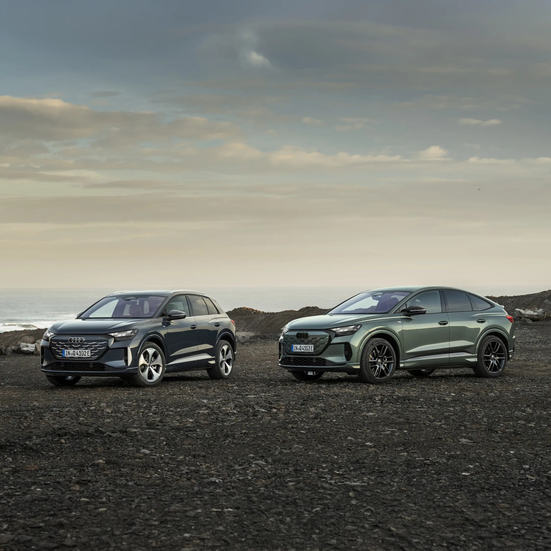 Two dark-colored Audi electric SUVs parked on rocky terrain with a cloudy sky in the background.